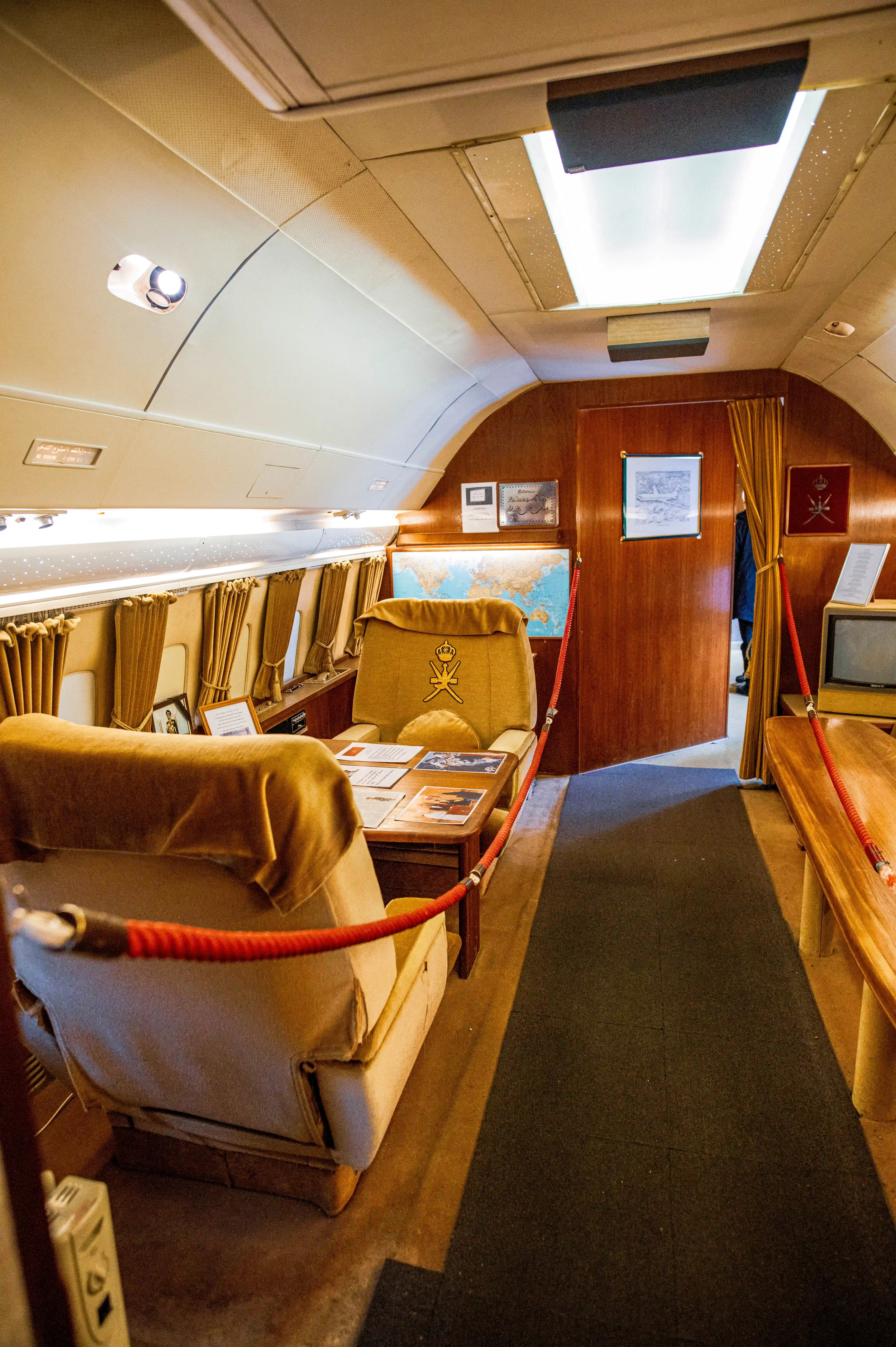 The interior of a private jet cabin, showing two large, comfortable chairs covered in tan fabric. One chair prominently displays a golden crest. The walls are panelled in dark wood, and a world map hangs on the wall.