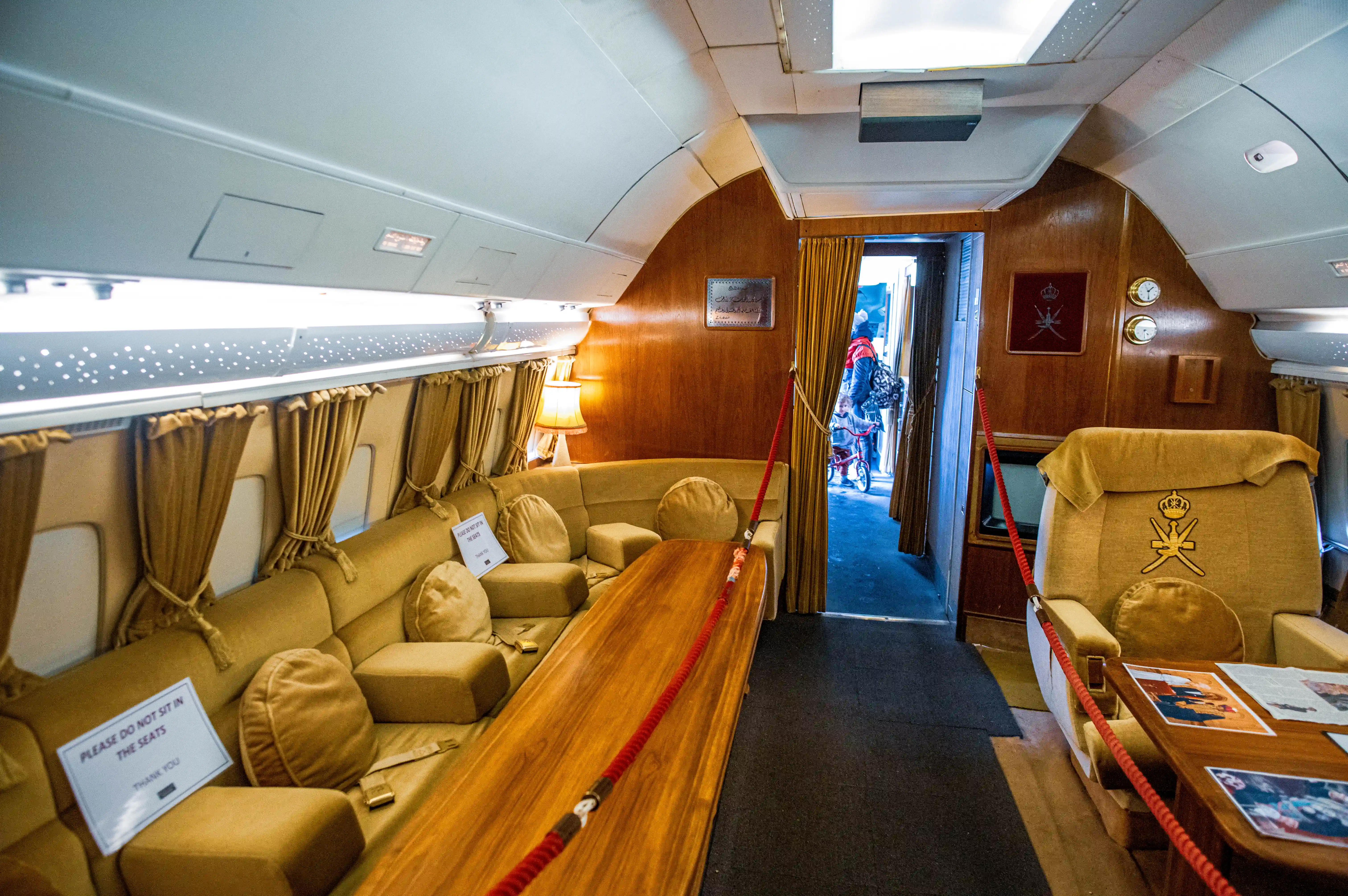 The interior of a vintage aircraft cabin decorated in gold and brown tones, with a long curved sofa on the left and a single armchair on the right. A wooden table sits in the centre, with red ropes preventing access.