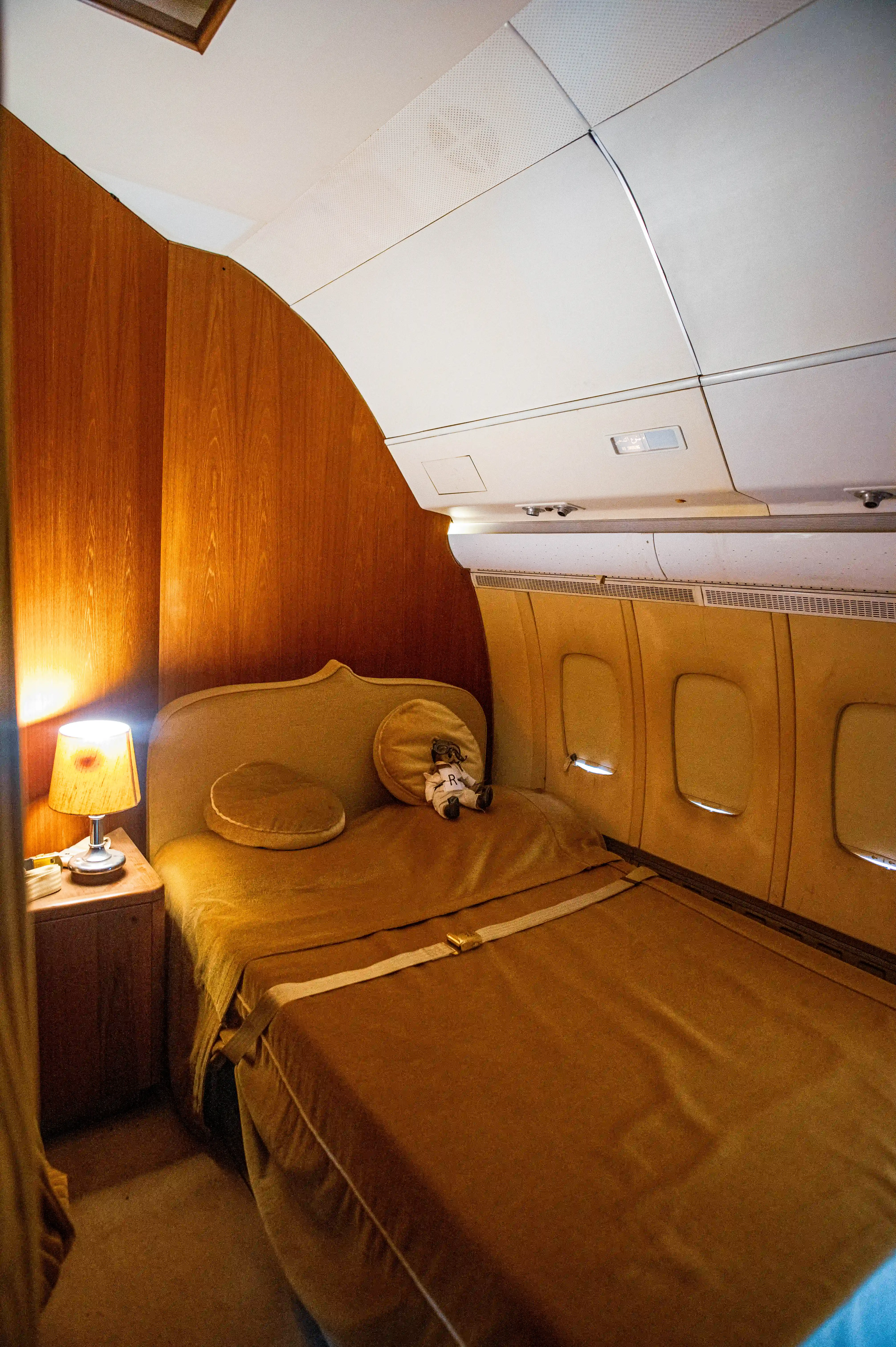 Interior view of a bedroom in a private jet, featuring wood panelling, a bed with golden-brown bedding and a seatbelt, a bedside table with a lamp, and small aircraft windows.