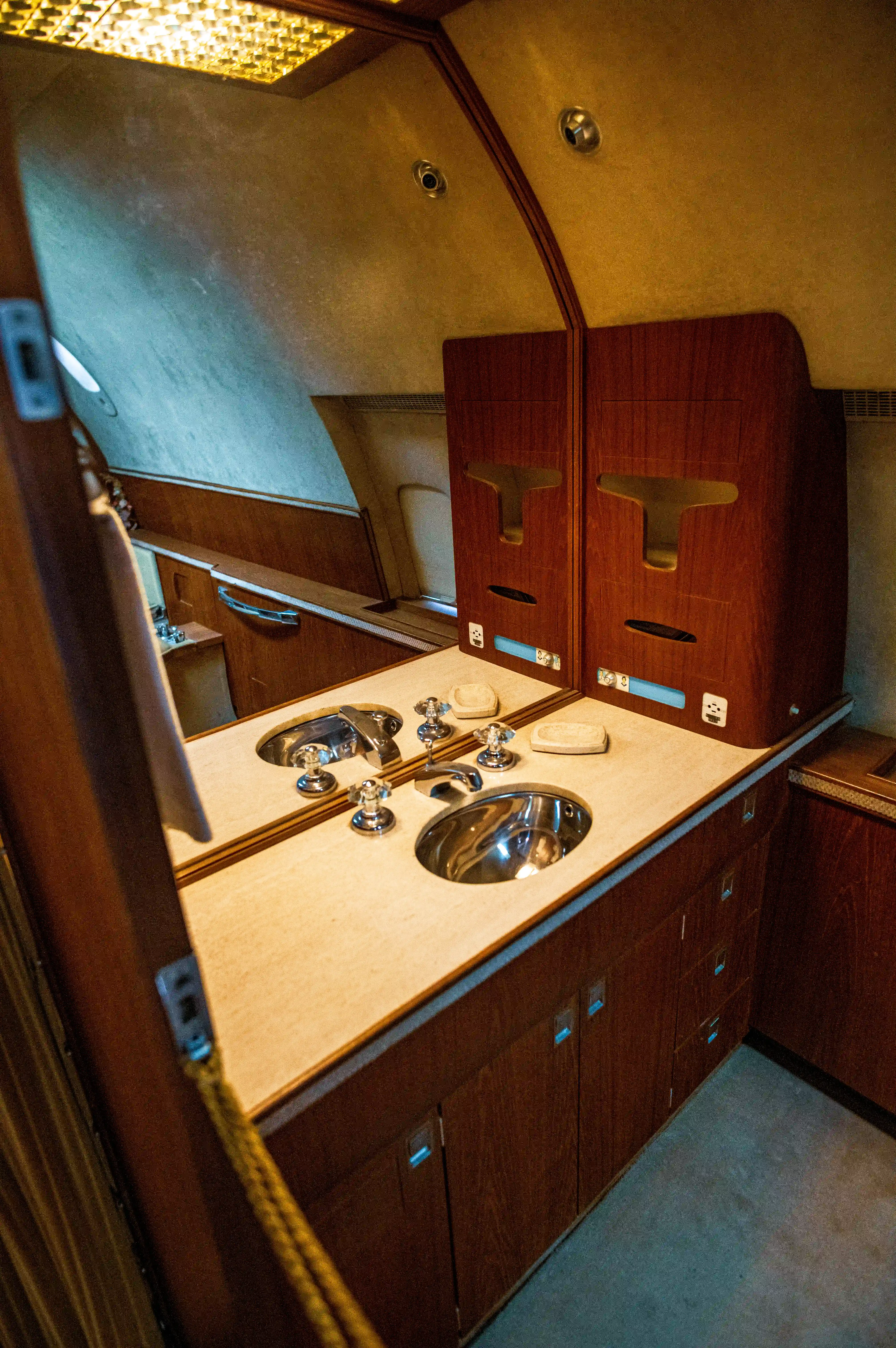 A view of a private jet washroom, showing a long vanity counter with two sinks, a large mirror reflecting the sinks and chrome taps, and dark wood cabinets below. The ceiling is textured and lit from above.