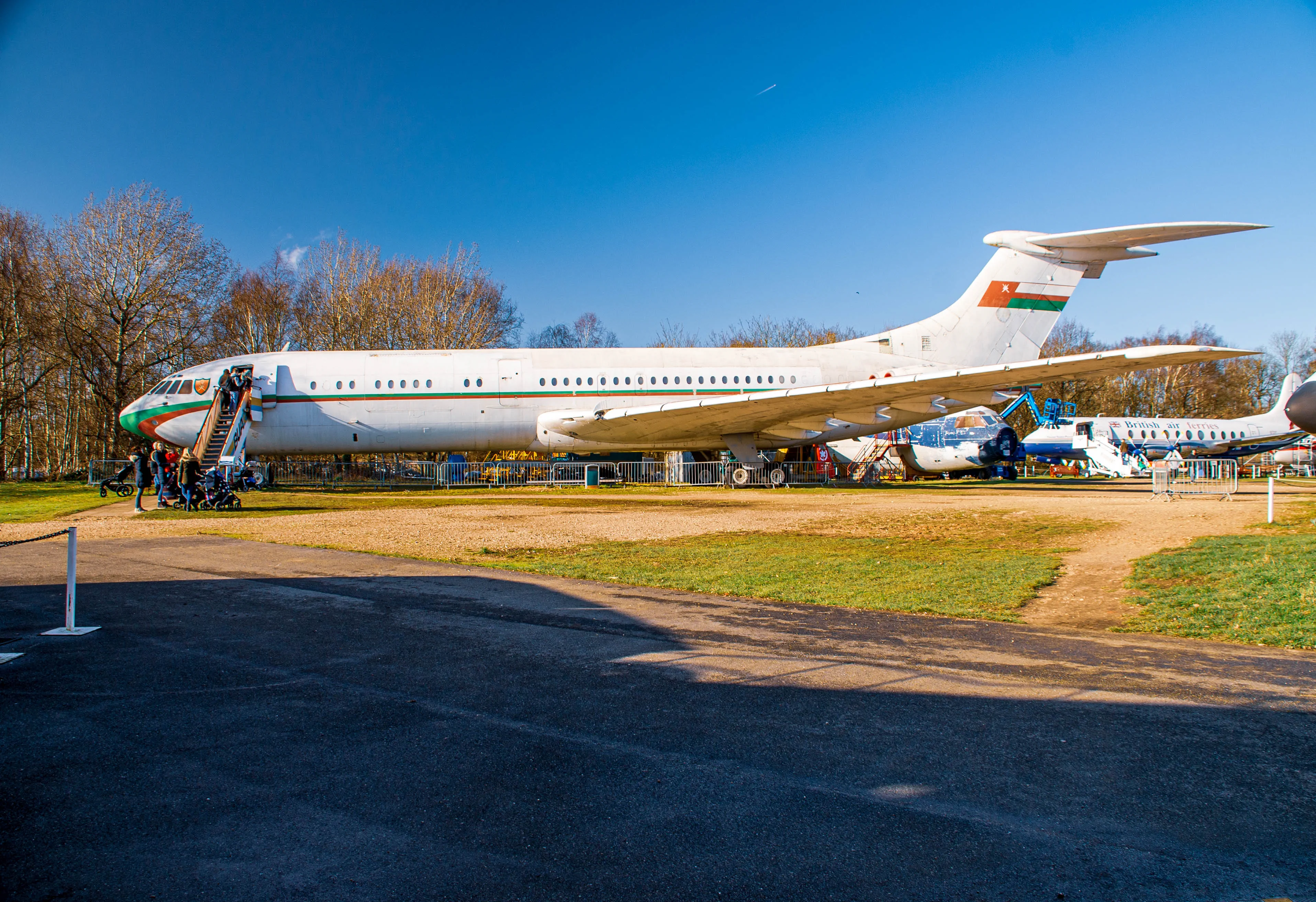 A large white vintage passenger jet aircraft with green and orange trim on the nose and a flag on its tail is parked on a grassy area. People are standing on the stairs leading into the aircraft. Another smaller aircraft with 
