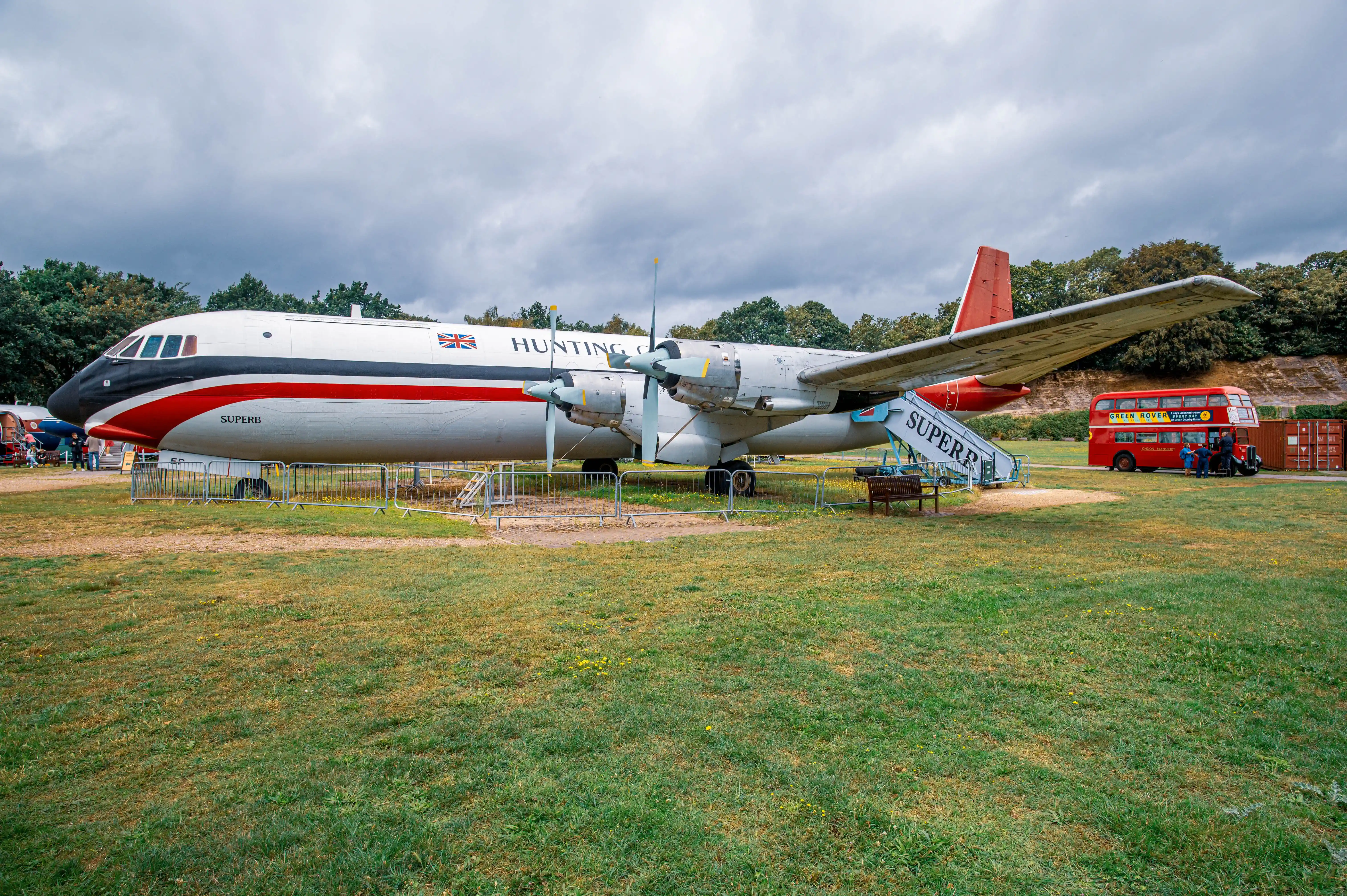  A large white and red airliner with HUNTING and SUPERB written on its fuselage, parked next to a red double-decker bus in a grassy field under a cloudy sky.