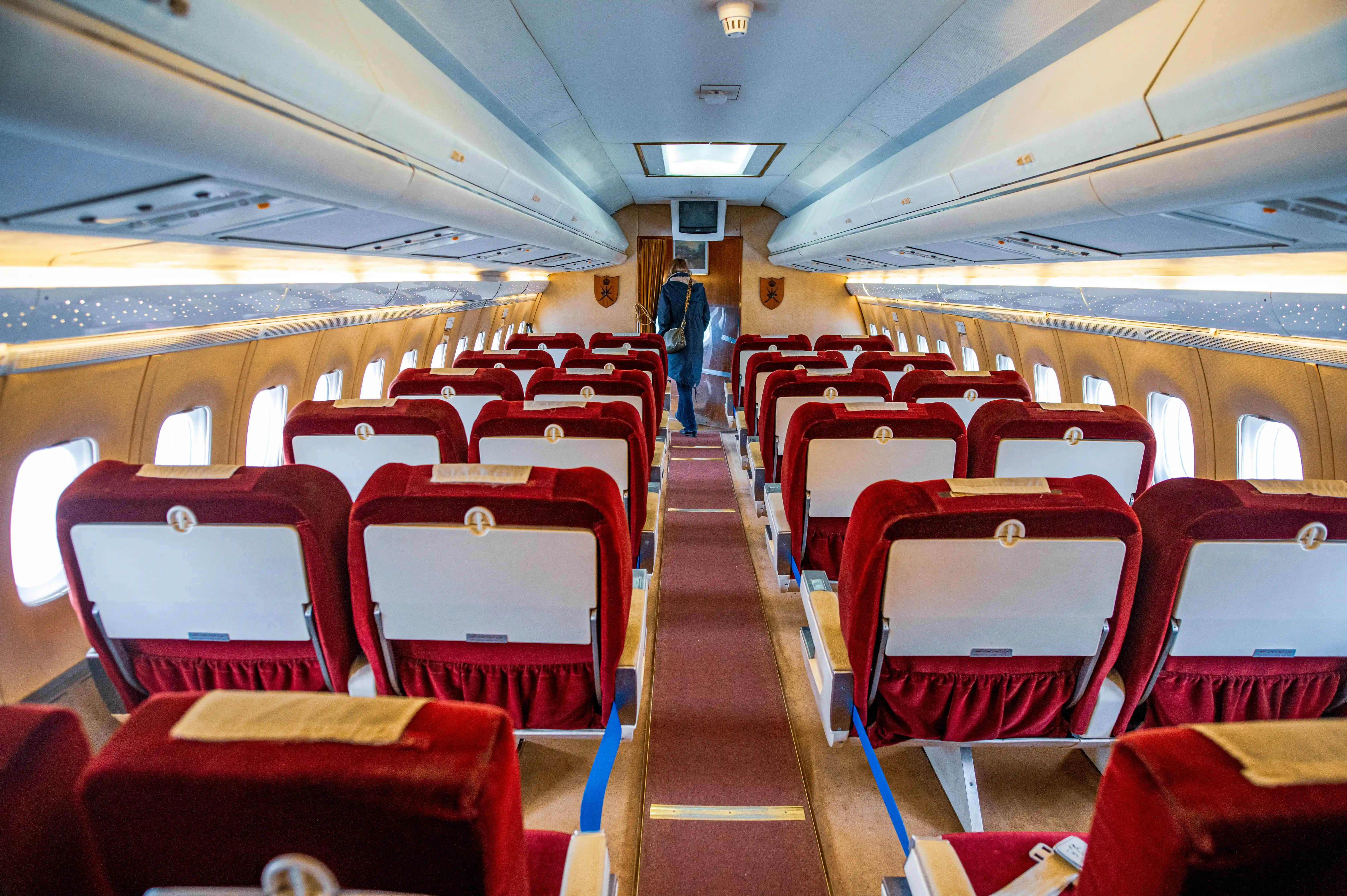 Interior view of an aeroplane cabin with numerous rows of deep red velvet seats, each with a white tray table. A person in a dark coat walks down the carpeted aisle towards the front of the plane, where a small screen and decorative elements are visible.