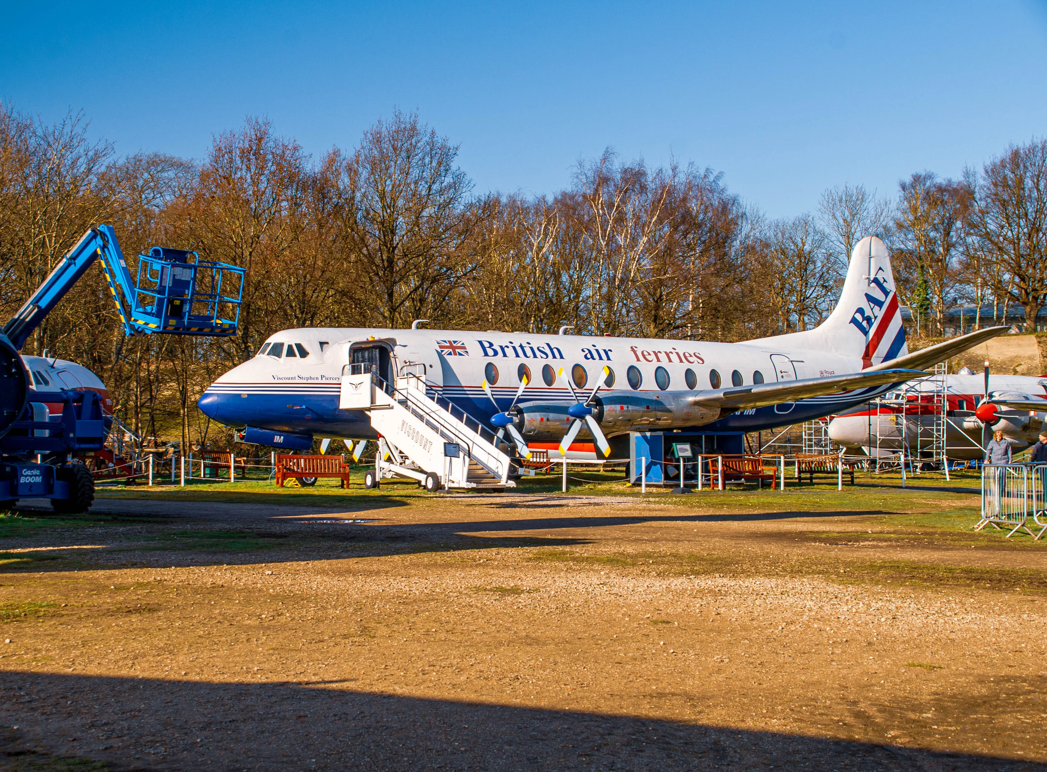 A white and blue twin-engine propeller aircraft with 'British air ferries' written on its side is parked outdoors. A blue scissor lift is to the left of the plane, and a set of stairs is deployed to the open cabin door. Two red benches are situated in front of the aircraft. In the background, bare trees and another aircraft under scaffolding are visible.