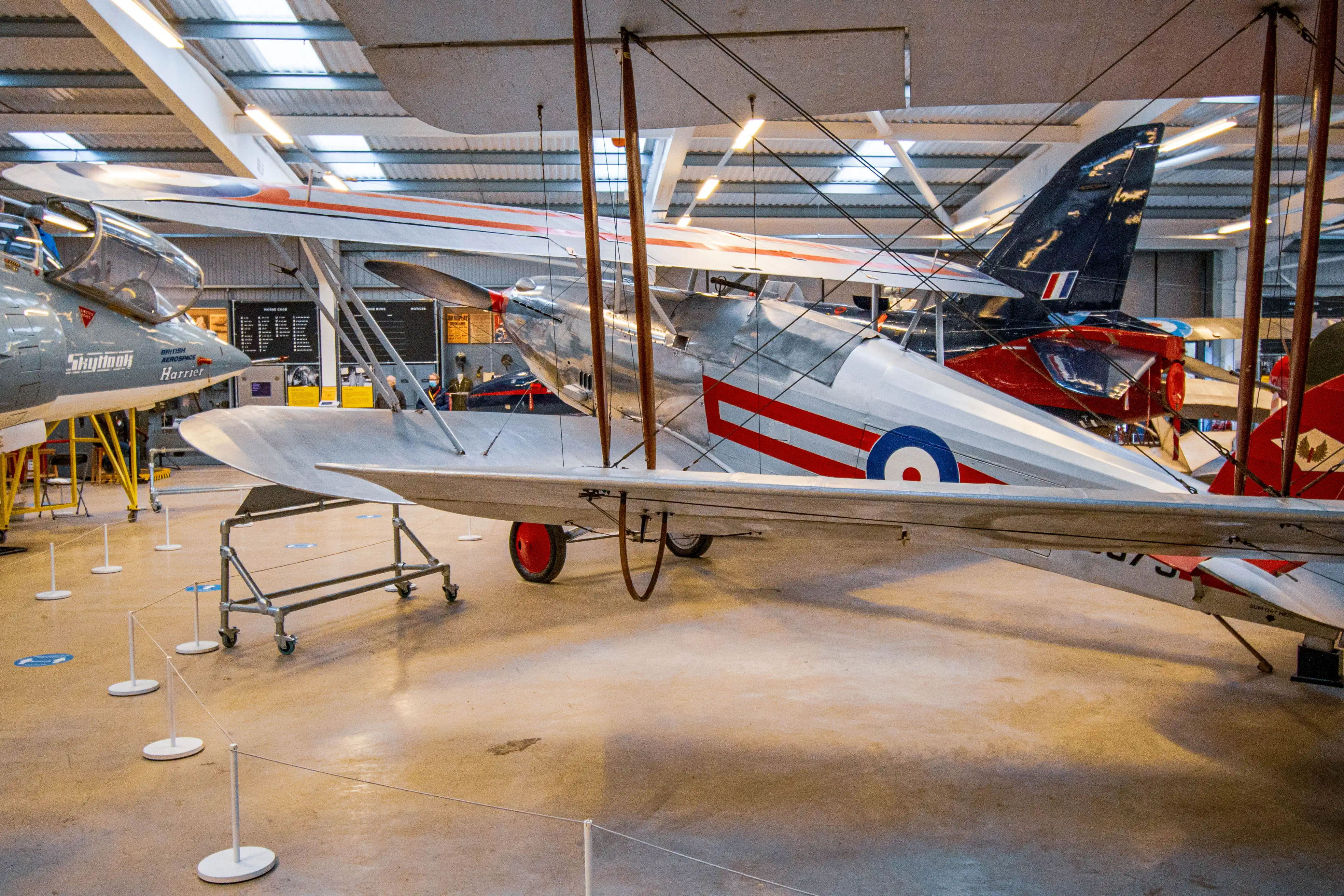 A vintage biplane with silver fuselage and red and blue markings is parked indoors. A modern grey jet fighter is to the left, and the tail of a red and blue aircraft is visible to the right.