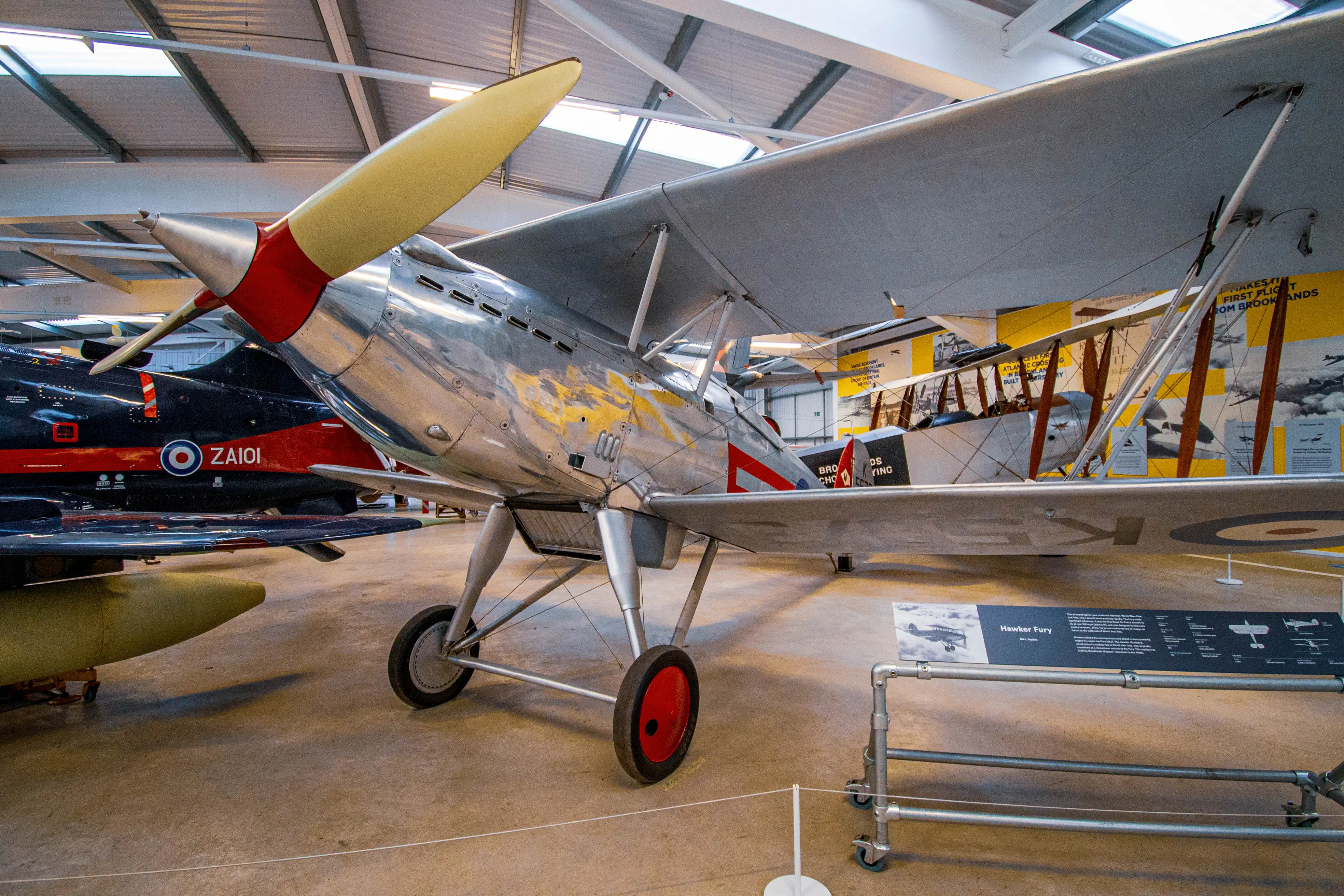A vintage silver biplane with a yellow-tipped propeller and red detailing sits on a museum floor, with a modern dark blue jet aircraft in the background.