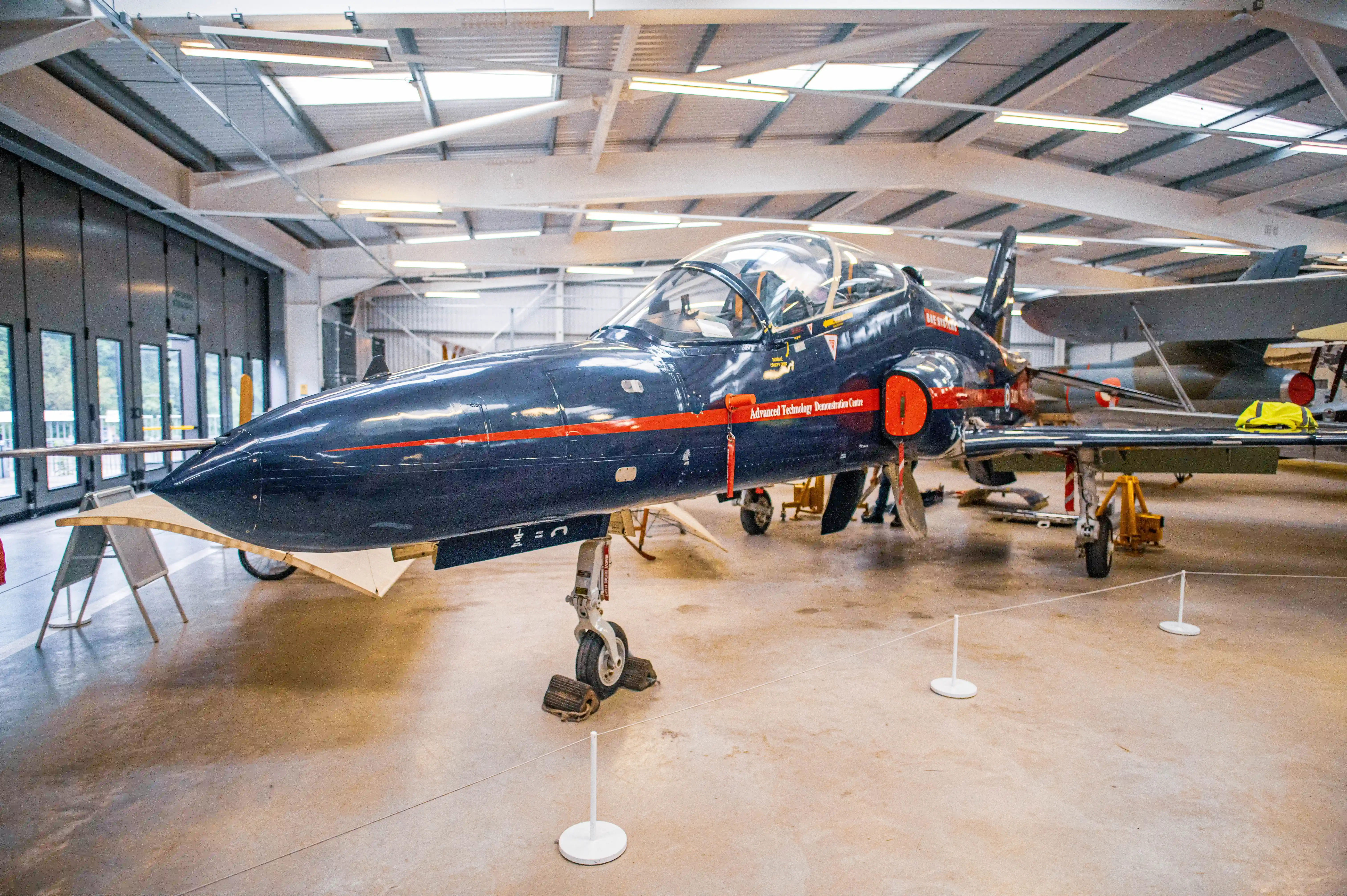 A dark blue jet aircraft with orange trim and the text Advanced Technology Demonstration Centre is parked inside a hangar.