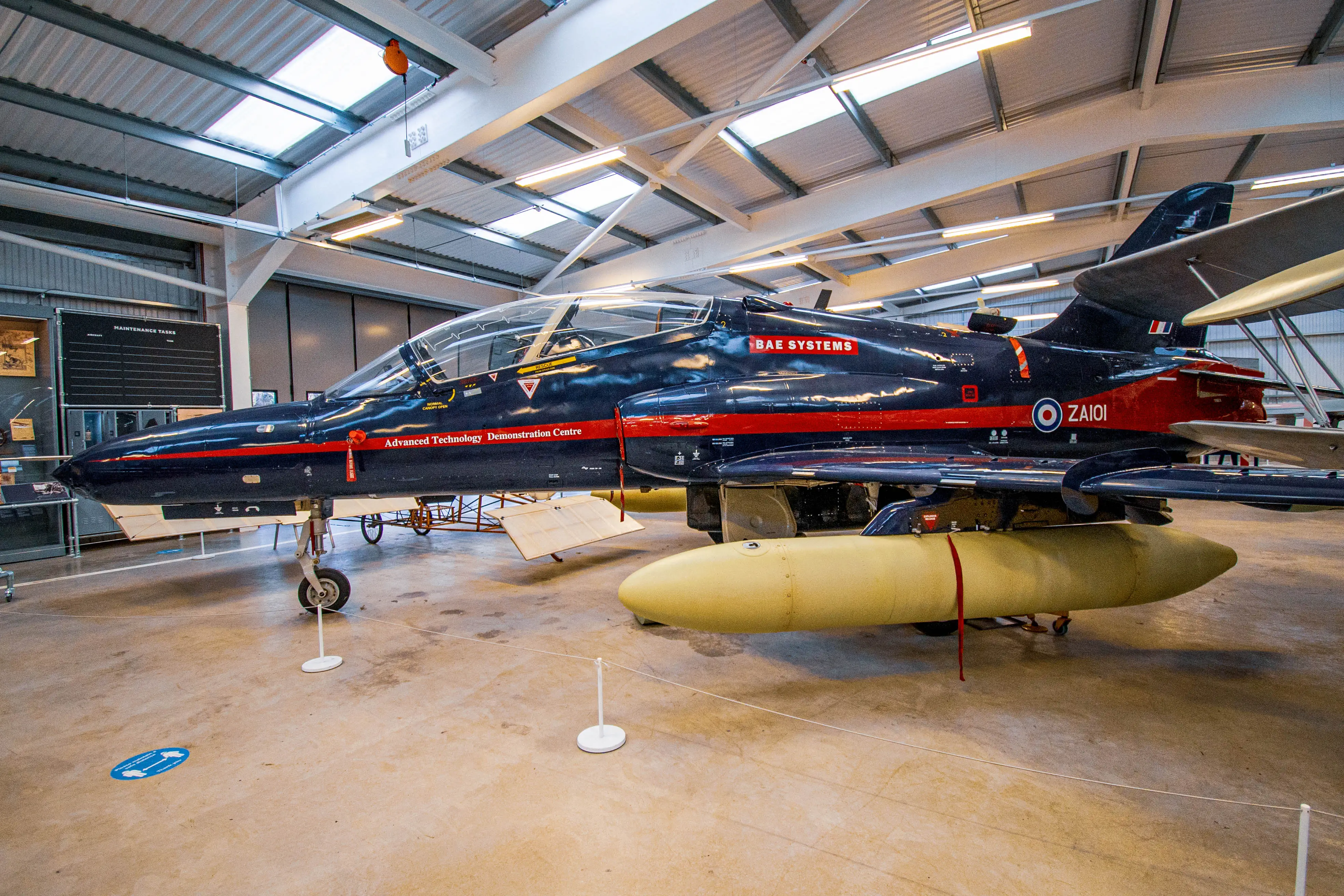 Side view of a dark blue and red BAE Systems Hawk jet aircraft with yellow auxiliary fuel tanks visible under the wings, parked inside a well-lit hangar. The aircraft is labelled ZAI01 and 