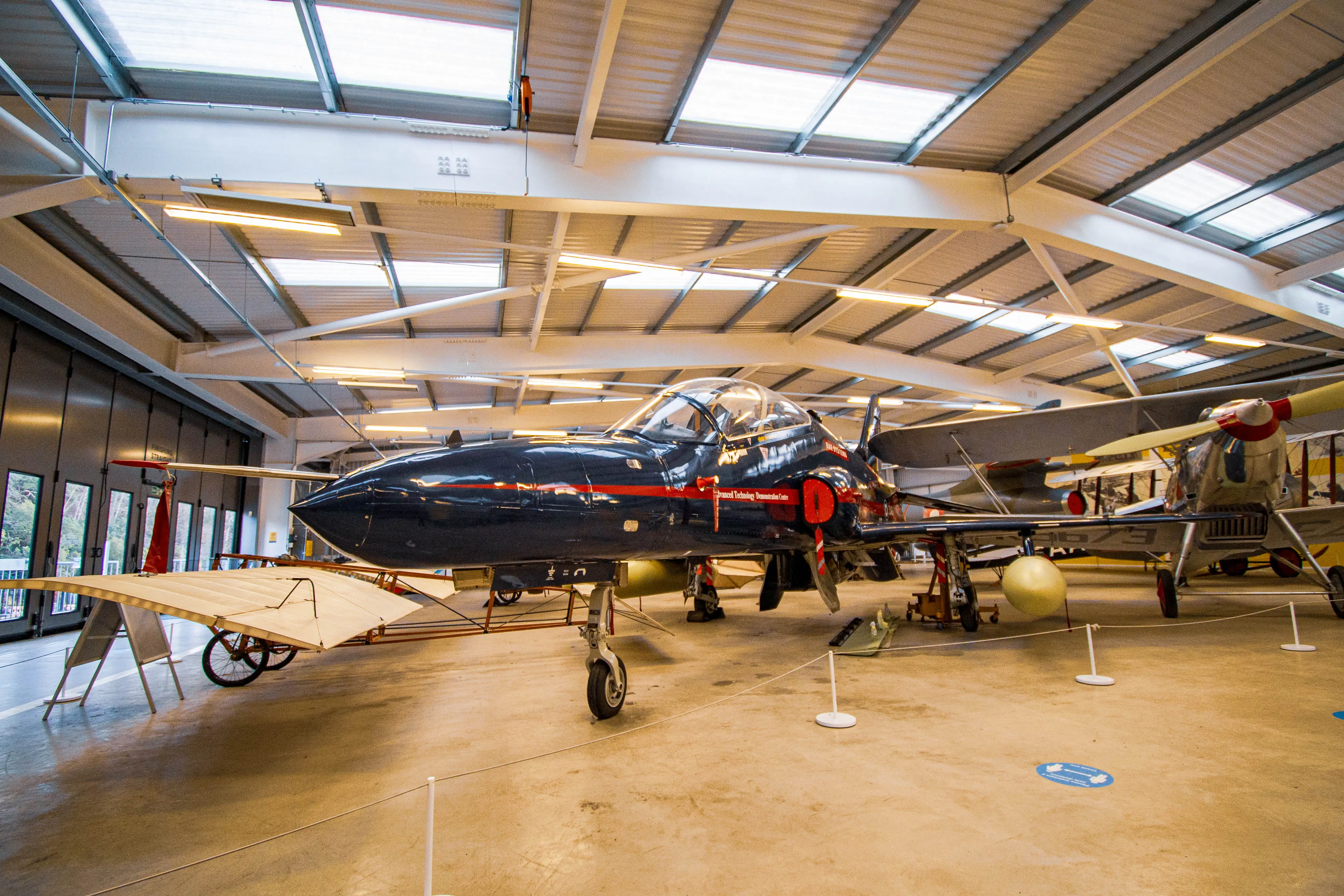 A dark blue jet aircraft with red accents is parked in a museum hangar. In the background, a biplane with a propeller is visible. The aircraft are displayed under a roof with skylights and fluorescent lighting.
