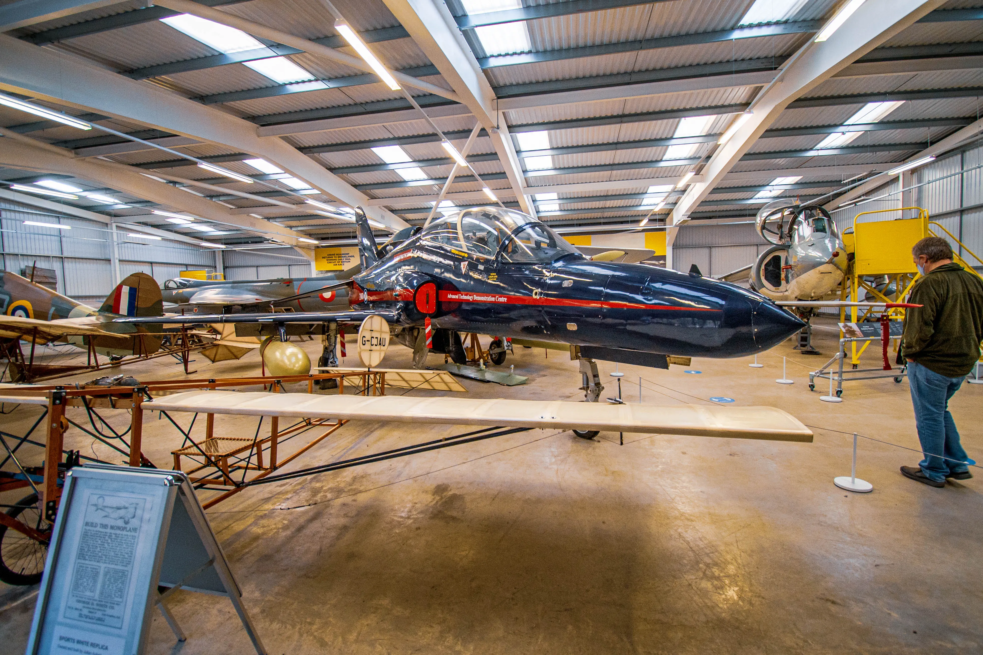 A museum hangar displays a collection of vintage aircraft, with a prominent dark blue jet in the foreground. A man stands to the right, viewing the aircraft.