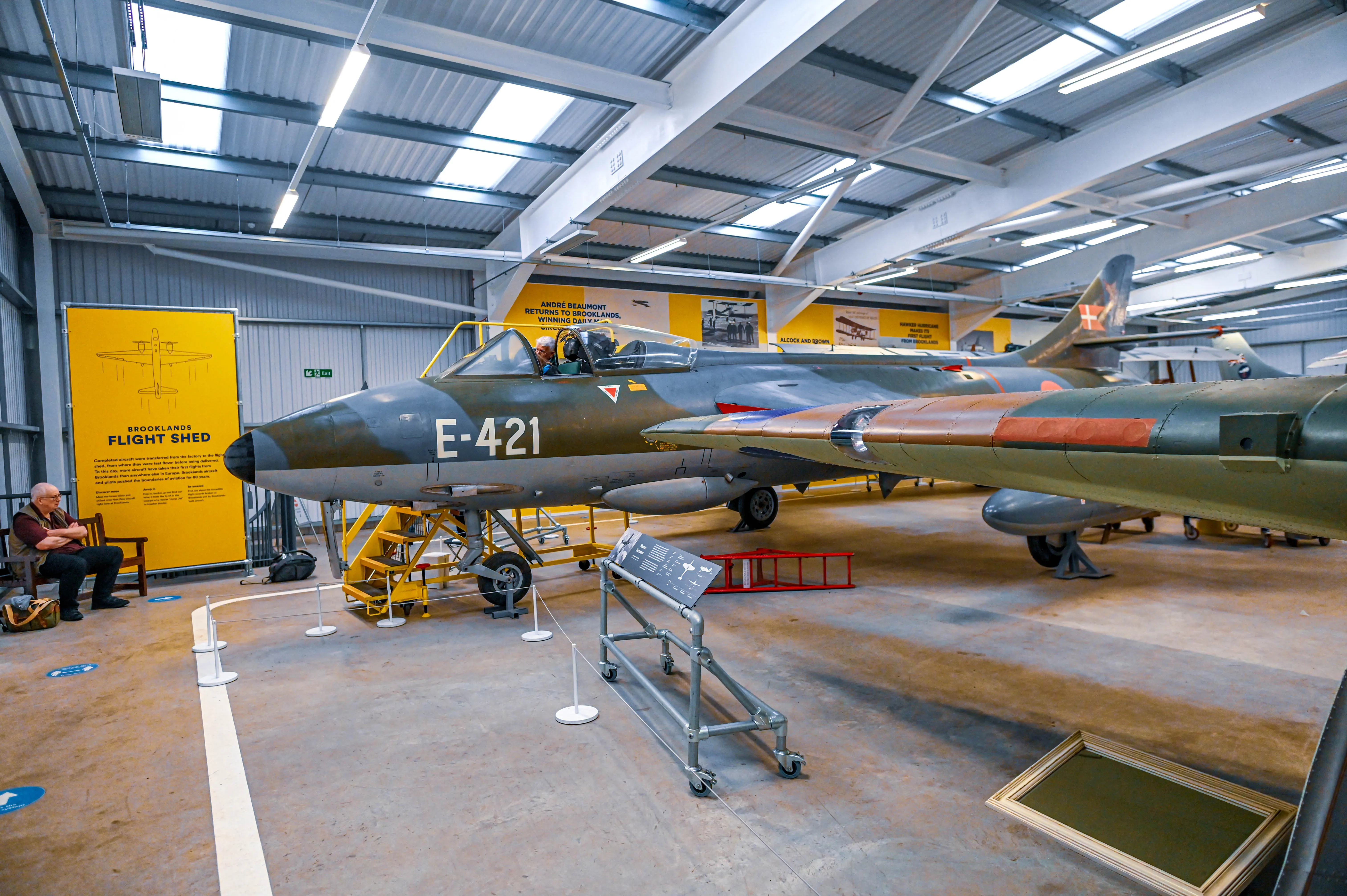 A close-up view of a vintage grey and green camouflage painted Hawker Hunter fighter jet aircraft, identified as E-421, parked inside a museum hangar. The aircraft's cockpit, wings, and landing gear are visible, along with a yellow access platform. Other exhibits and aircraft parts are in the background under a large, corrugated metal roof with skylights.