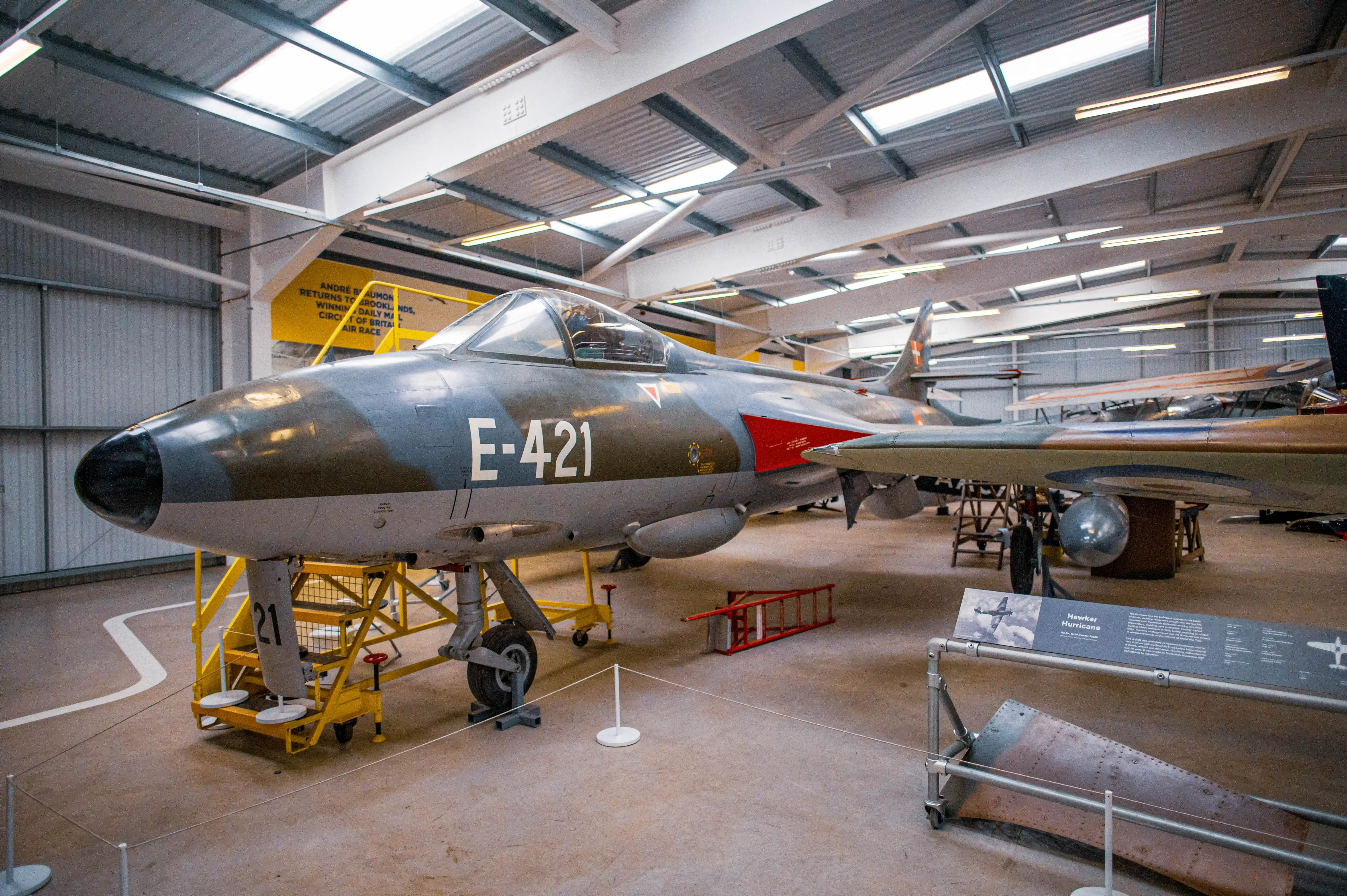 A close-up view of a vintage grey and green camouflage painted Hawker Hunter fighter jet aircraft, identified as E-421, parked inside a museum hangar. The aircraft's cockpit, wings, and landing gear are visible, along with a yellow access platform. Other exhibits and aircraft parts are in the background under a large, corrugated metal roof with skylights.