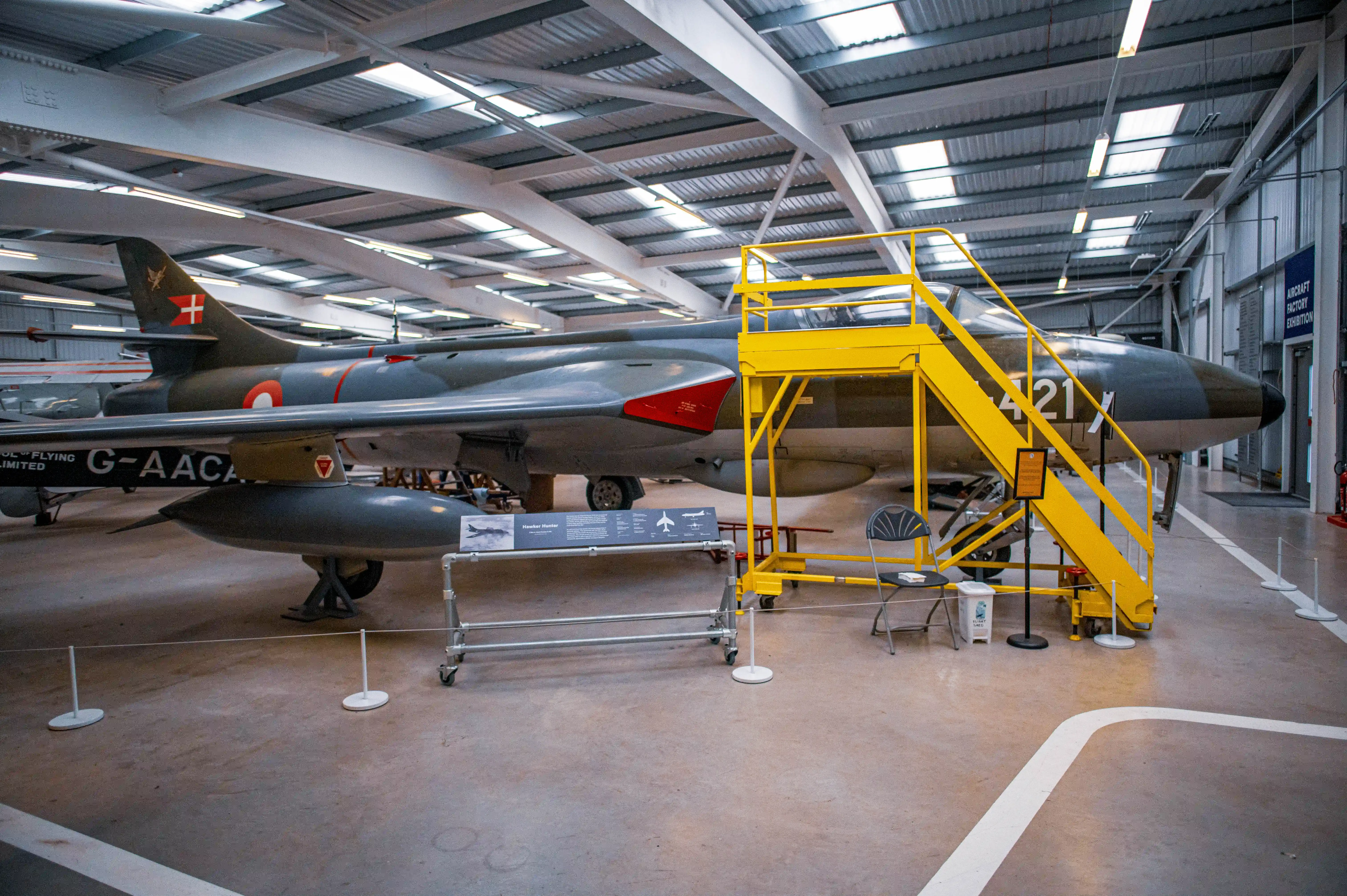 Side view of a dark green Hawker Hunter fighter jet with red accents, parked inside a museum. A bright yellow set of stairs leads up to the cockpit.