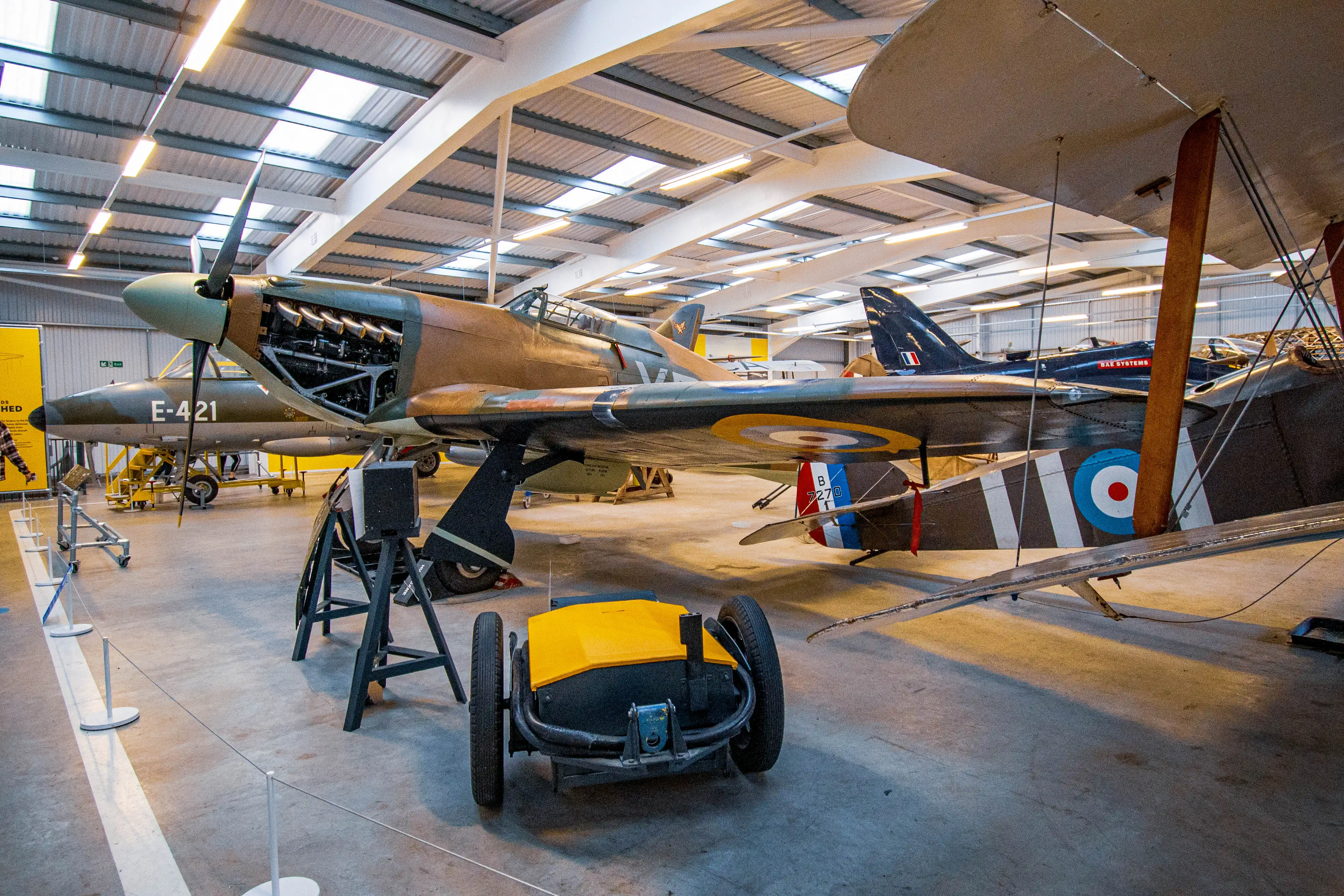 Interior view of a museum displaying several vintage aircraft, including a Hawker Hurricane and a biplane, with a yellow ground handling tug in the foreground.
