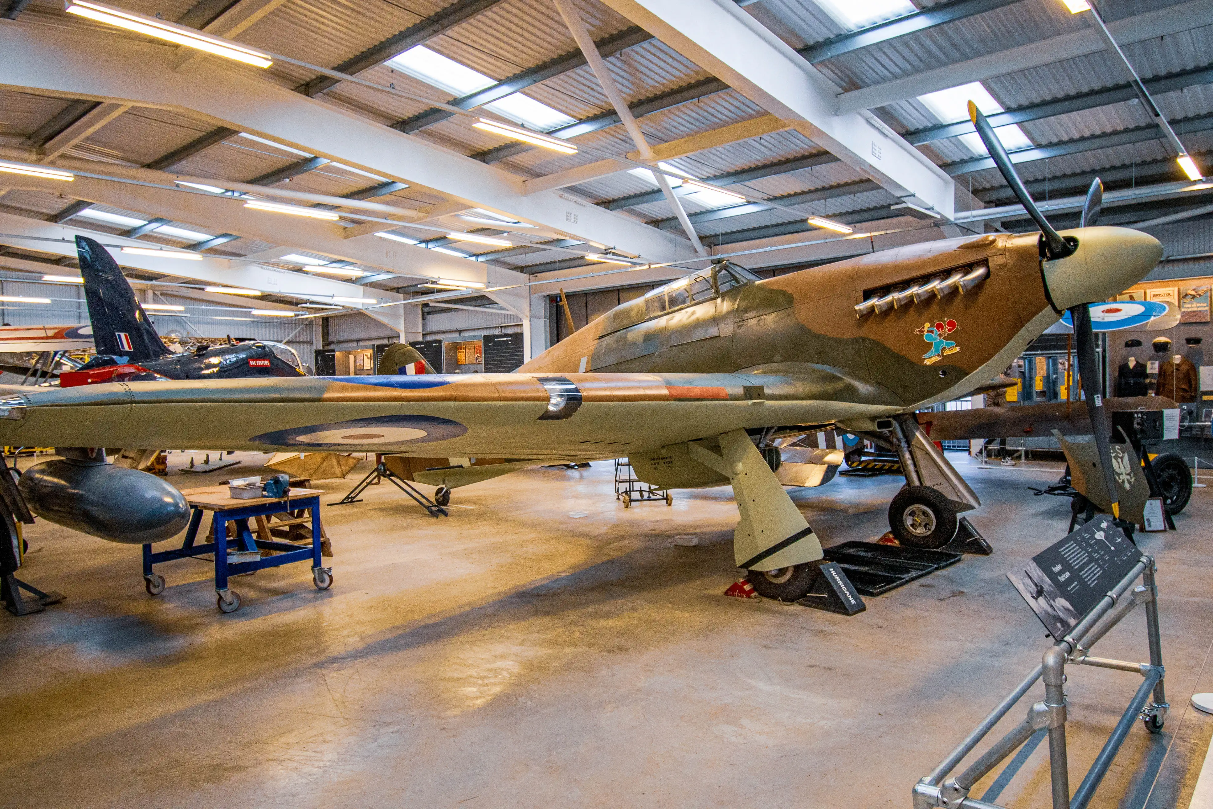 A vintage military aircraft with a camouflage-painted fuselage and RAF roundel markings displayed inside a modern hangar. The aircraft features a propeller, machine gun ports, and military insignia on its body. The hangar is well-lit with industrial lighting, and other aircraft and equipment are visible in the background