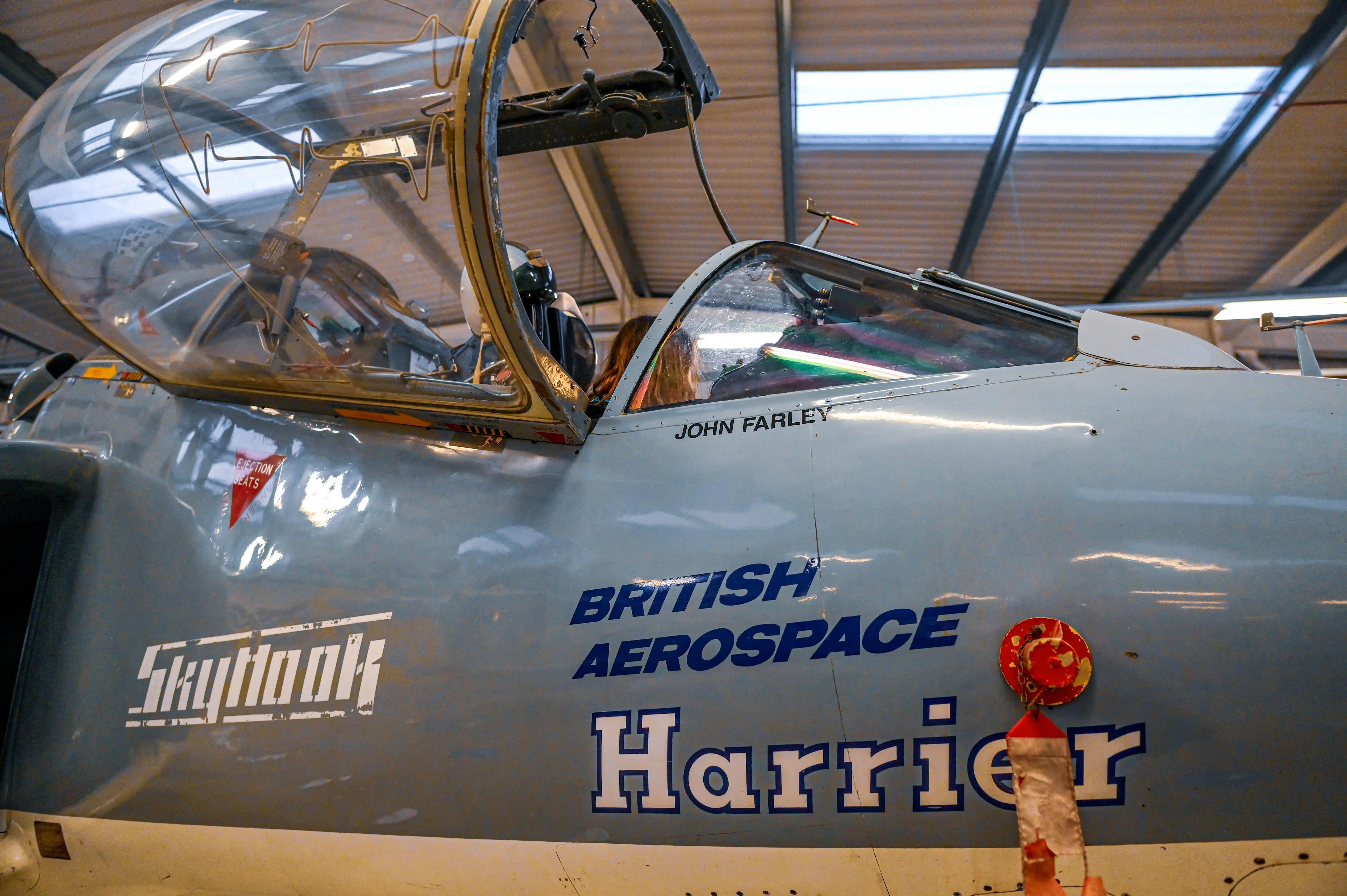 Close-up view of the cockpit of a light blue British Aerospace Harrier jet. The canopy is open, revealing part of the pilot's seat and controls. The words 'Skyhook' and 'BRITISH AEROSPACE Harrier' are visible on the side of the aircraft, along with the name 'JOHN FARLEY' and a red 'EJECTION SEATS' warning triangle.