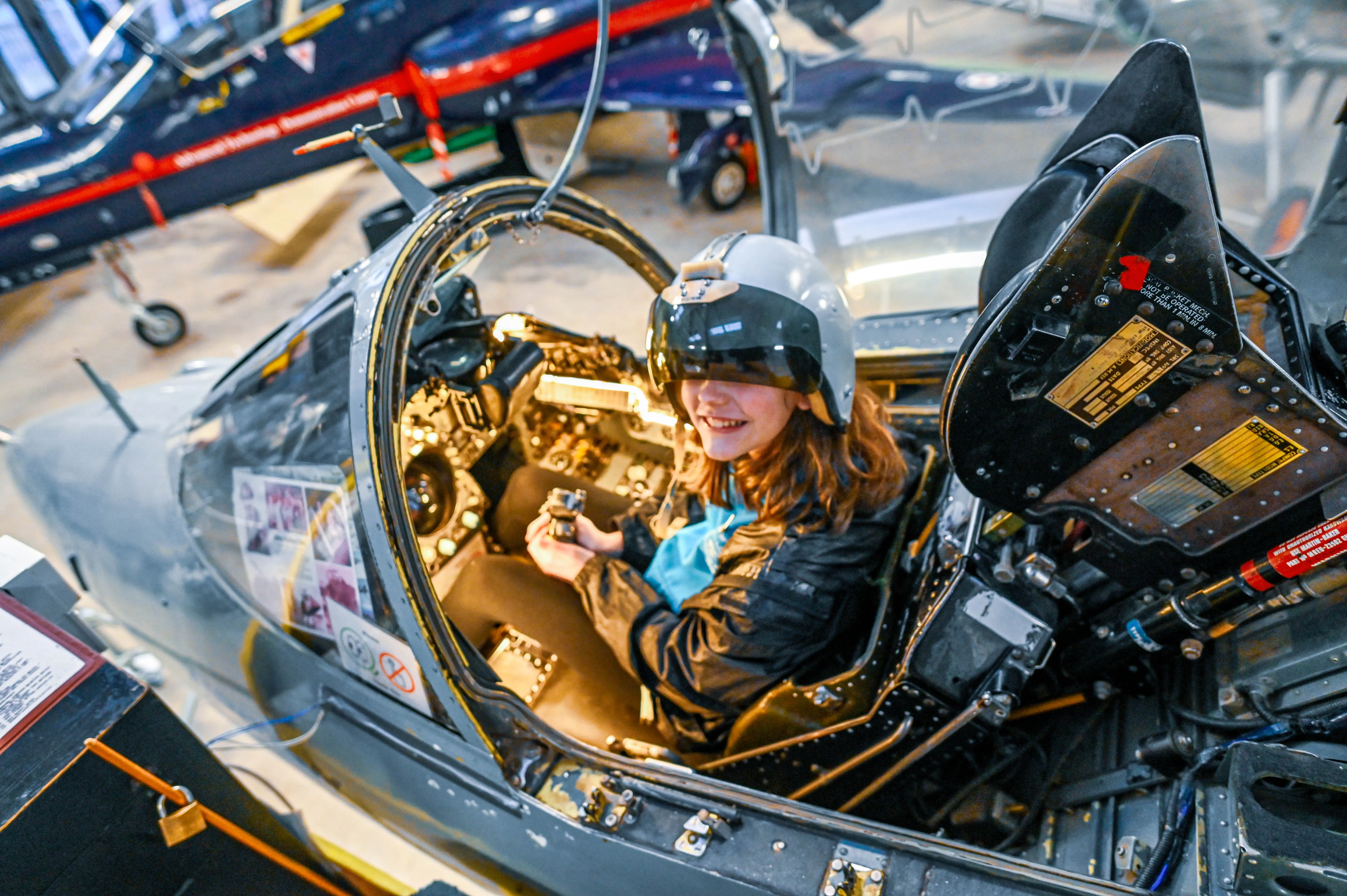 A young girl with a wide smile sits in the cockpit of a jet fighter, wearing a pilot's helmet and dark flight jacket. She holds the aircraft's controls in her hands.