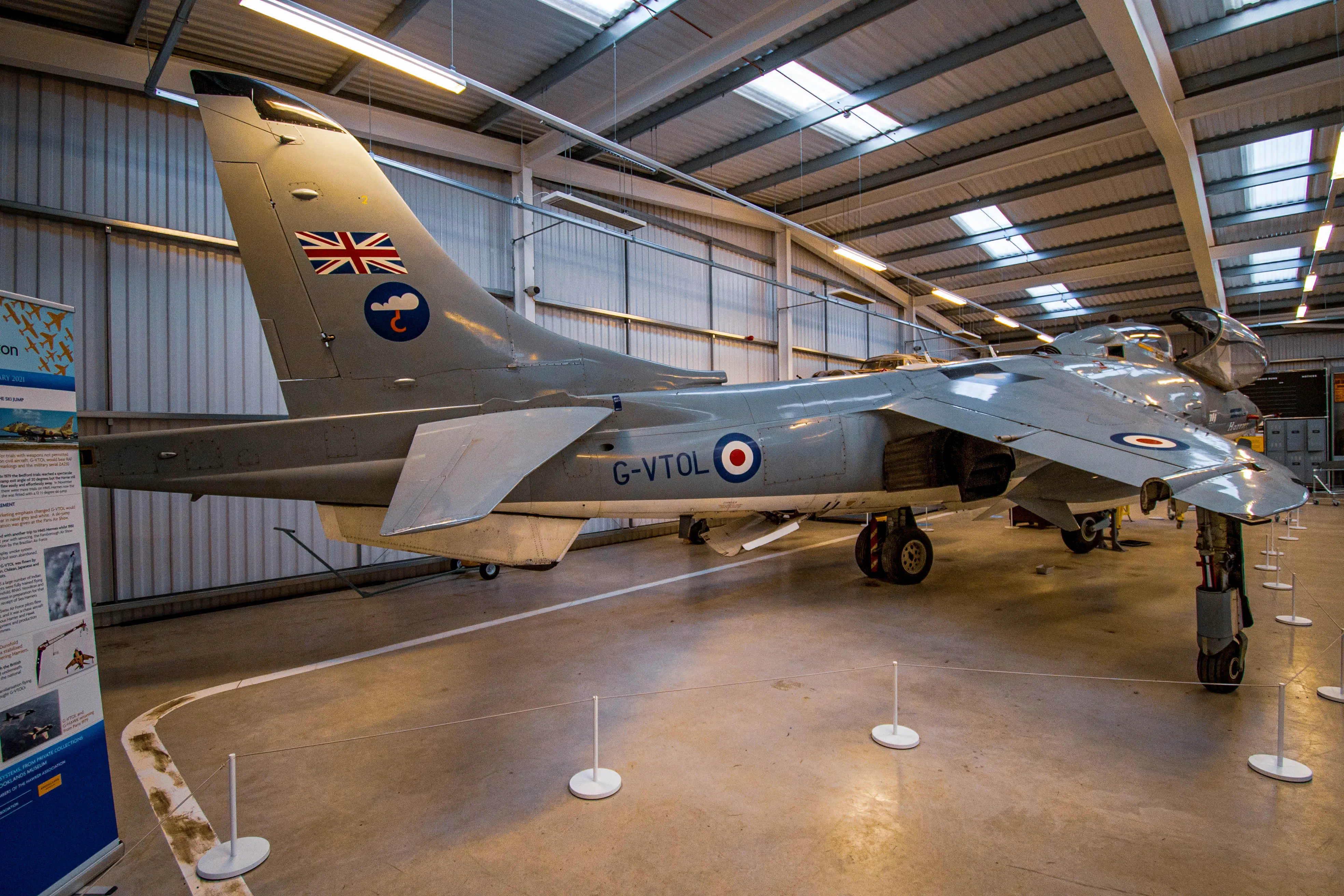 A rear three-quarter view of the Hawker Siddeley Harrier aircraft with the registration G-VTOL visible on its fuselage. The aircraft is displayed indoors, likely in a museum, with its distinctive grey livery and British roundels. The Union Jack and a cloud insignia are present on the tail fin.
