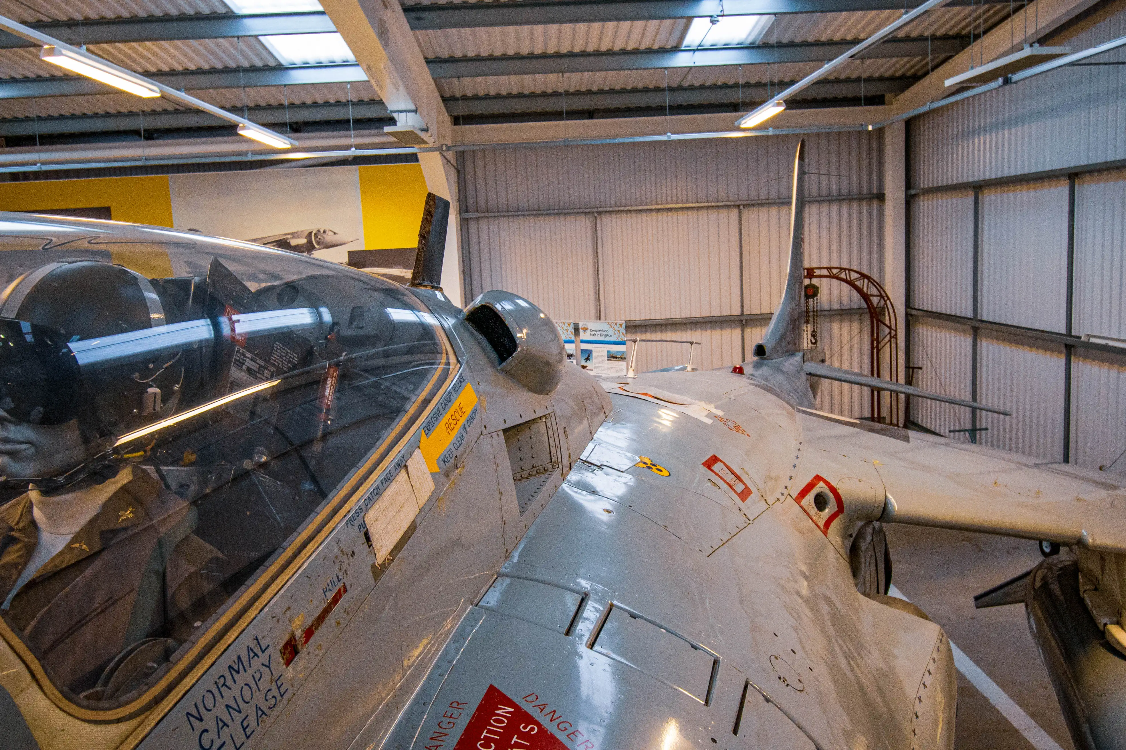 Close-up view of the cockpit of a Harrier jet on display, showing the pilot's seat, canopy, and warning labels on the grey exterior of the aircraft.