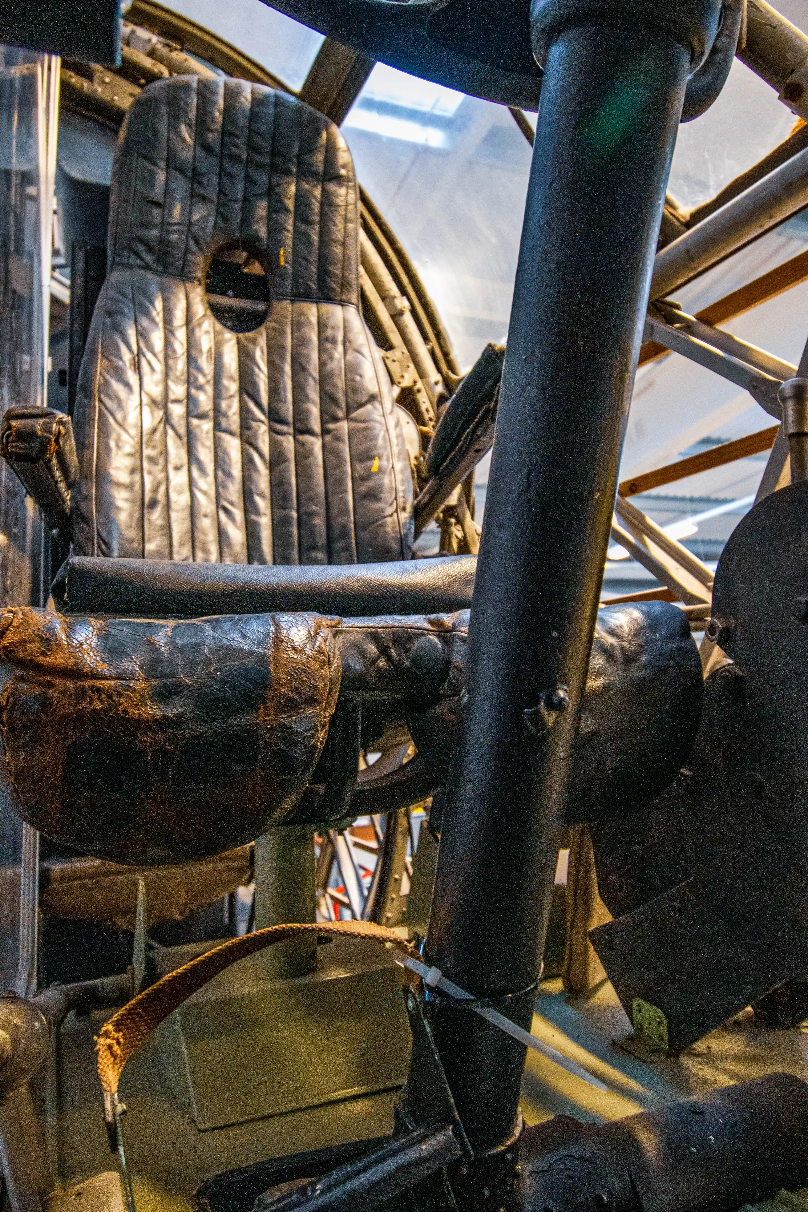 Interior view of an old aircraft cockpit showing a black leather pilot's seat with quilted padding and a circular headrest opening. A dark metal support column is prominent in the foreground, with visible structural elements of the aircraft in the background.