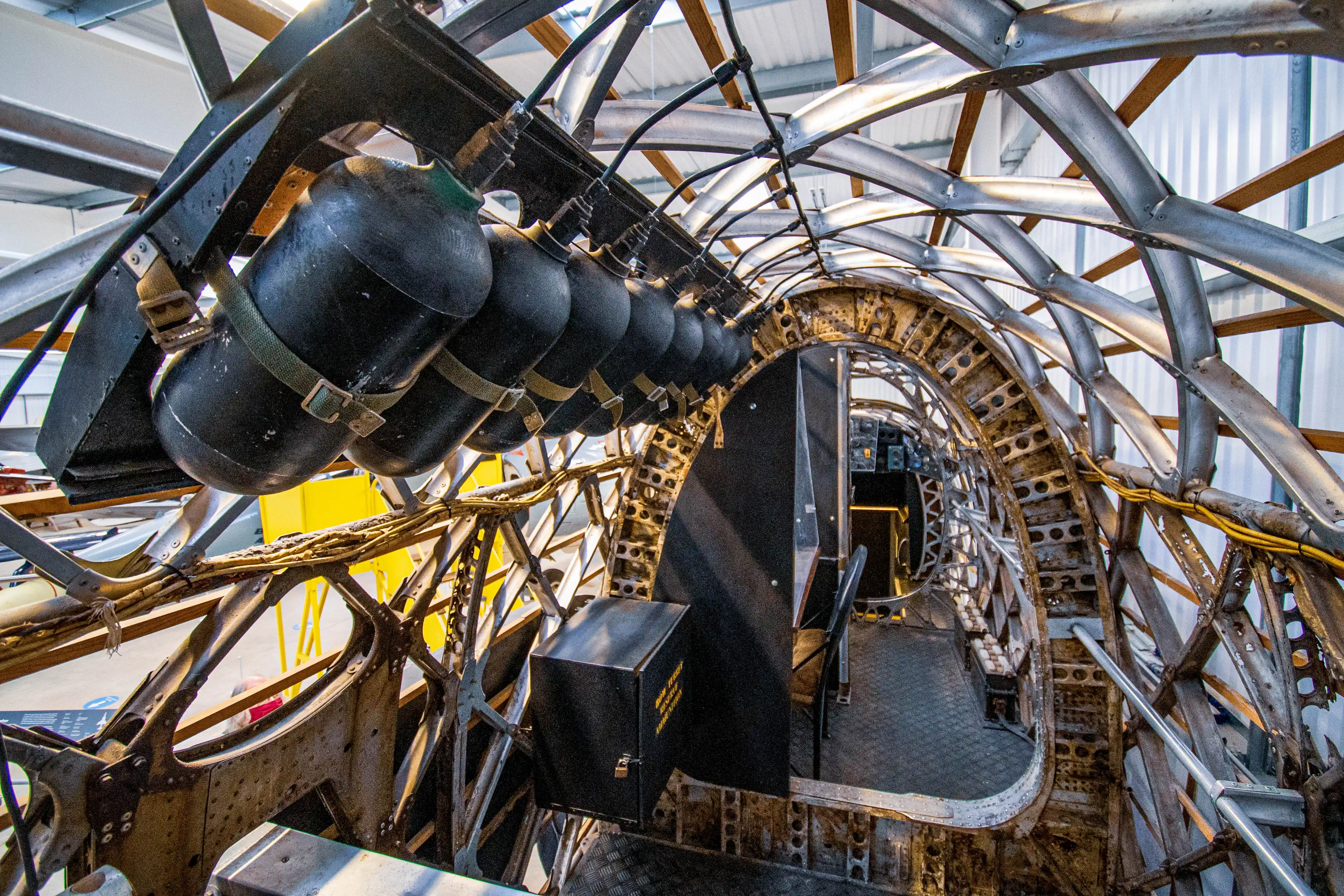 Inside the bare metal fuselage of a vintage aircraft, showing the exposed internal ribbing and framework, with a bank of black oxygen cylinders hanging overhead to the left.