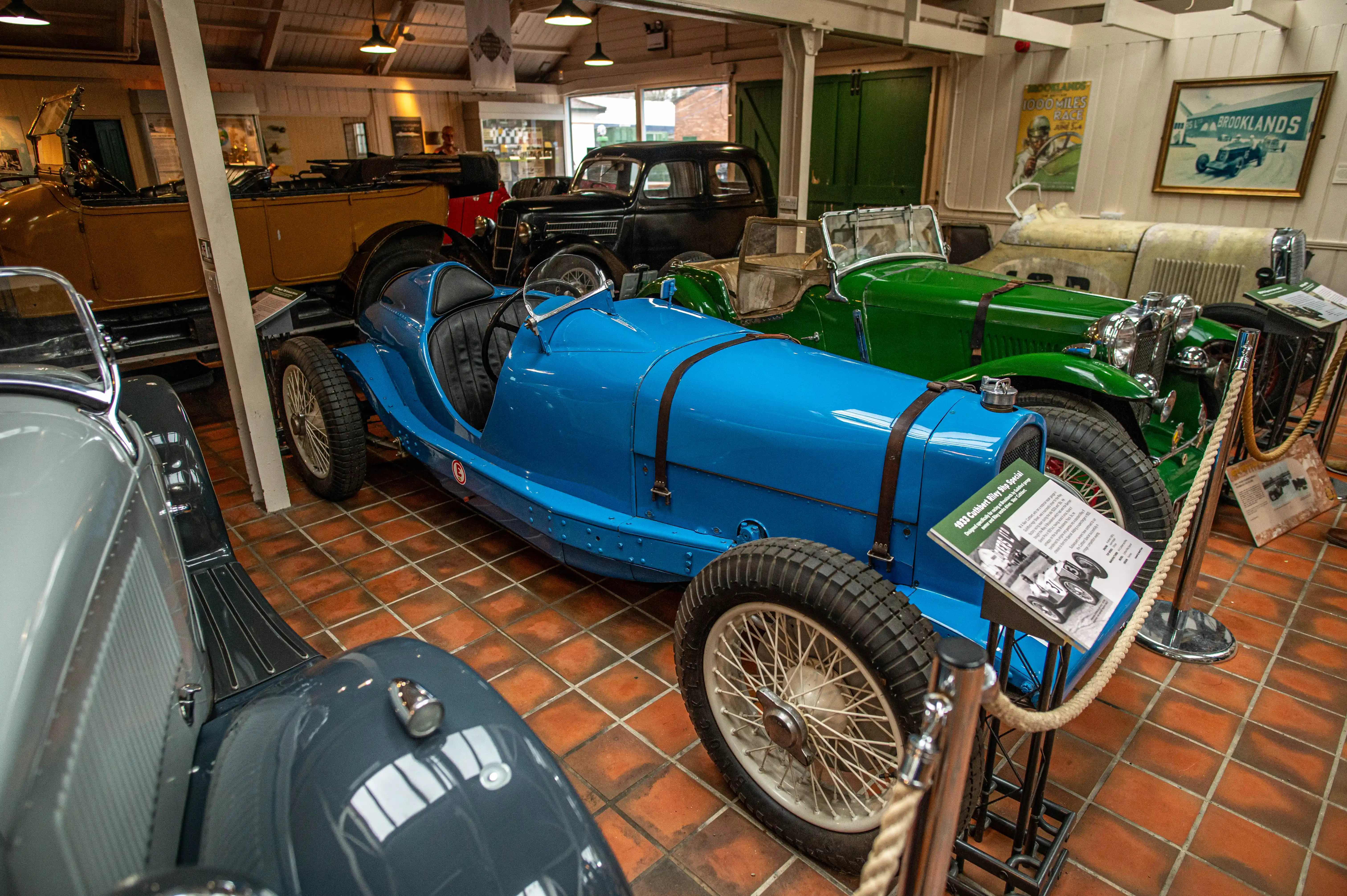 A vibrant blue 1933 single-seater racing car with wire-spoke wheels is prominently displayed in a museum, with a green vintage car and a black saloon car visible behind it.