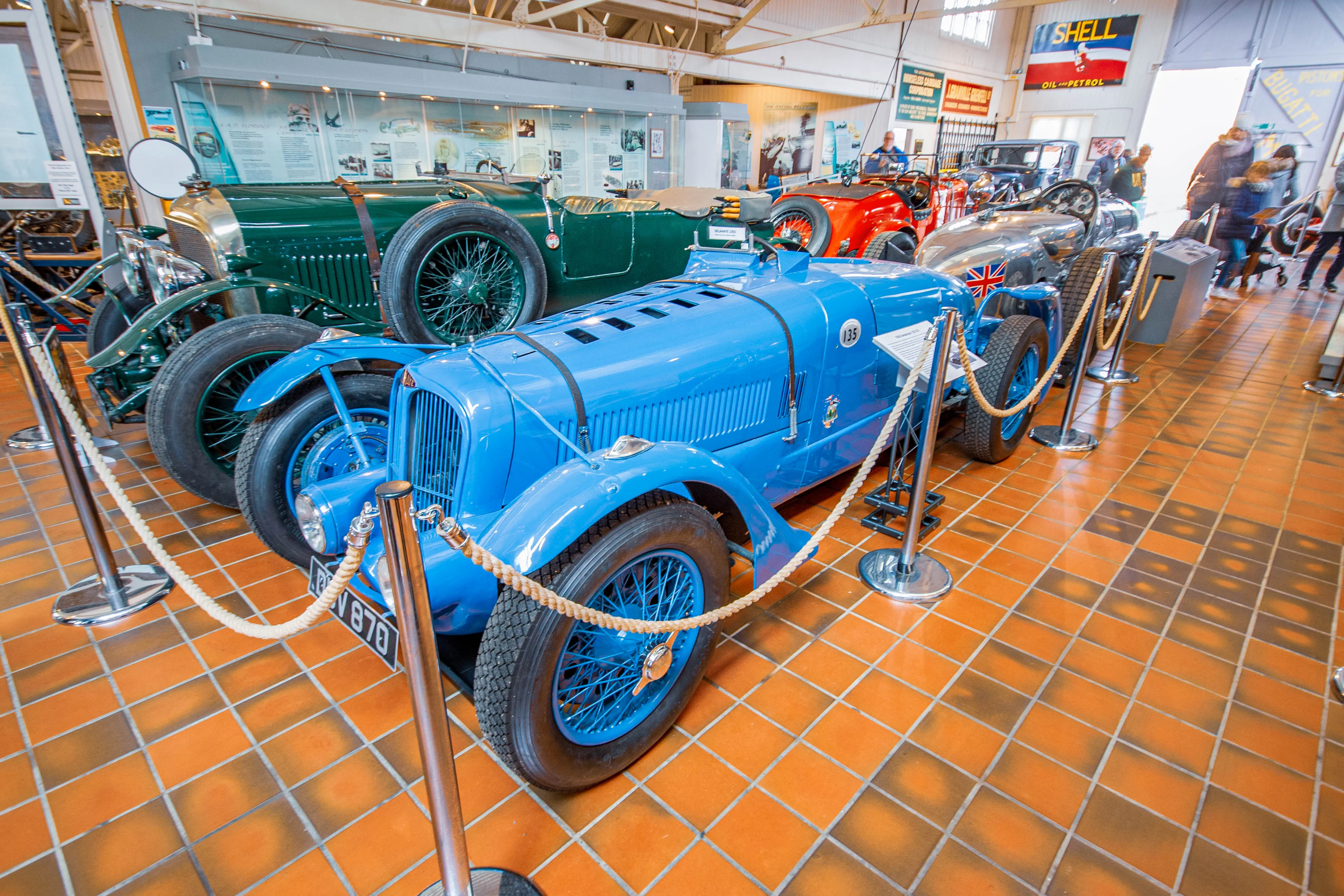 A bright blue vintage racing car with wire-spoke wheels and the number 135 on its side is displayed indoors on a tiled floor, with other vintage cars, including a green one and a silver one, visible in the background.