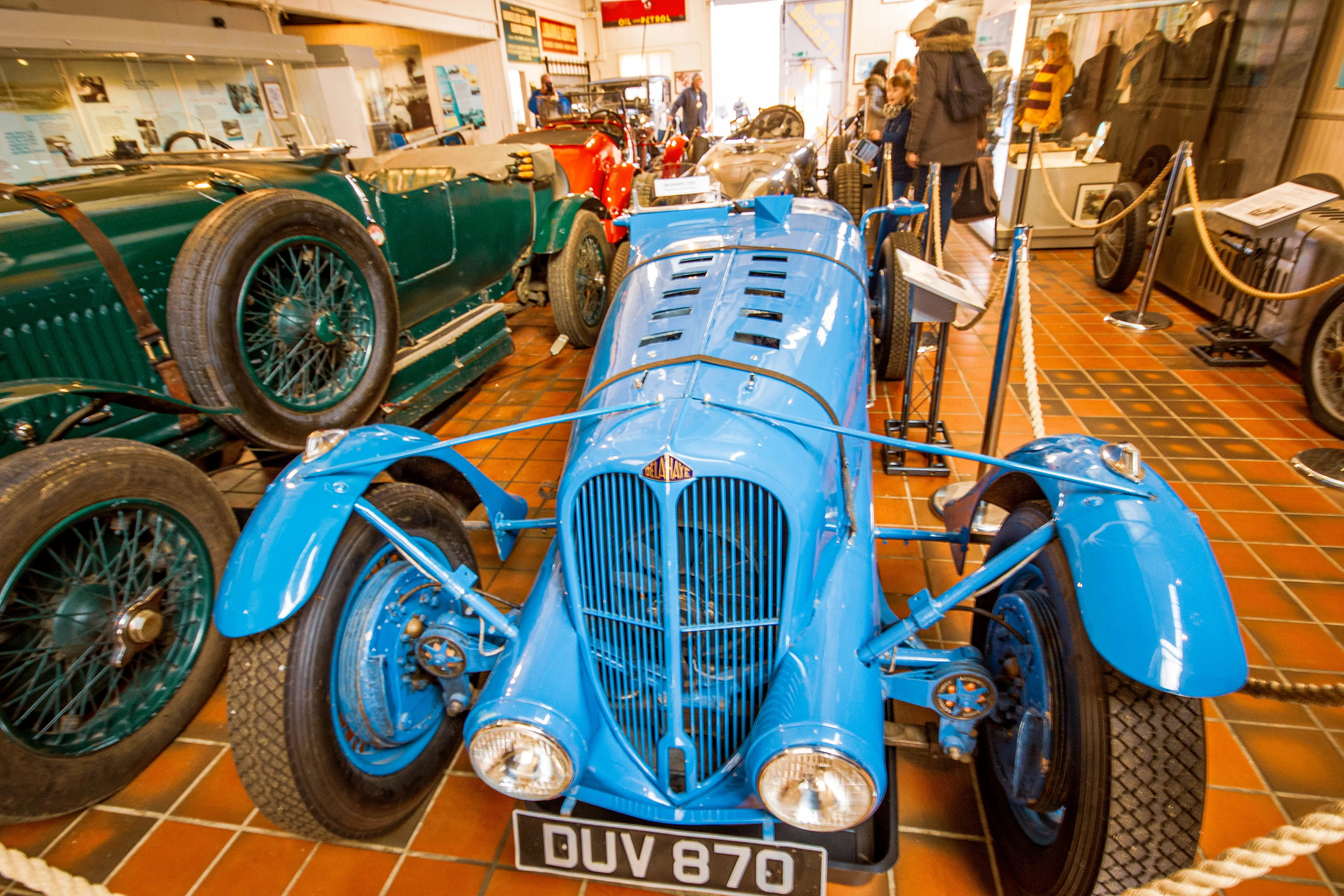 A bright blue vintage racing car with the license plate DUV 870 is parked in a museum, with a green vintage car to its left. People can be seen in the background looking at the cars.