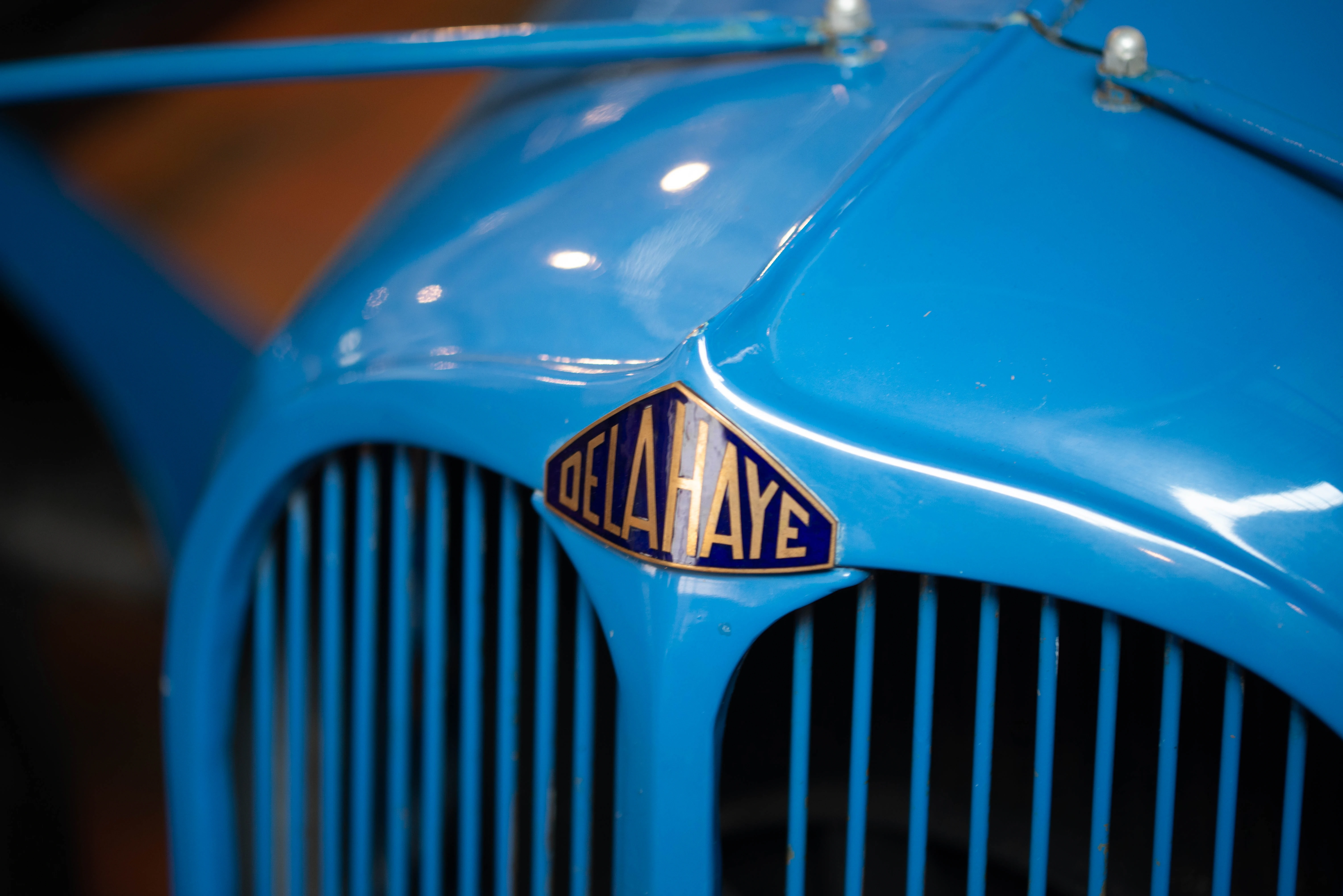 Close-up of a bright blue vintage car grille with a hexagonal Delahaye badge featuring gold lettering on a blue background.