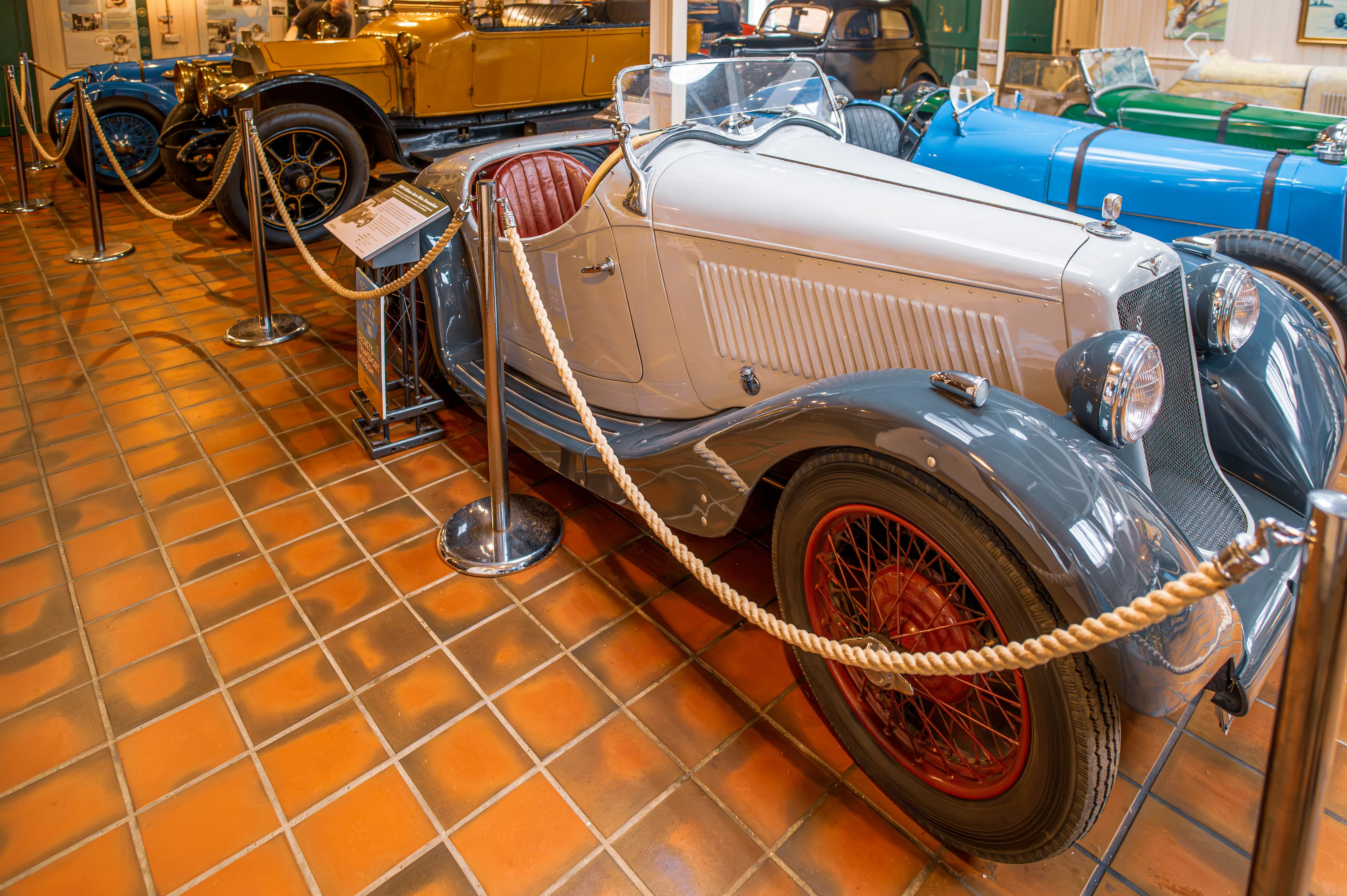 Interior view of a car museum with several vintage automobiles displayed, featuring a grey and white two-seater in the foreground, with other cars in blue, yellow, and green behind it, all separated by rope barriers.
