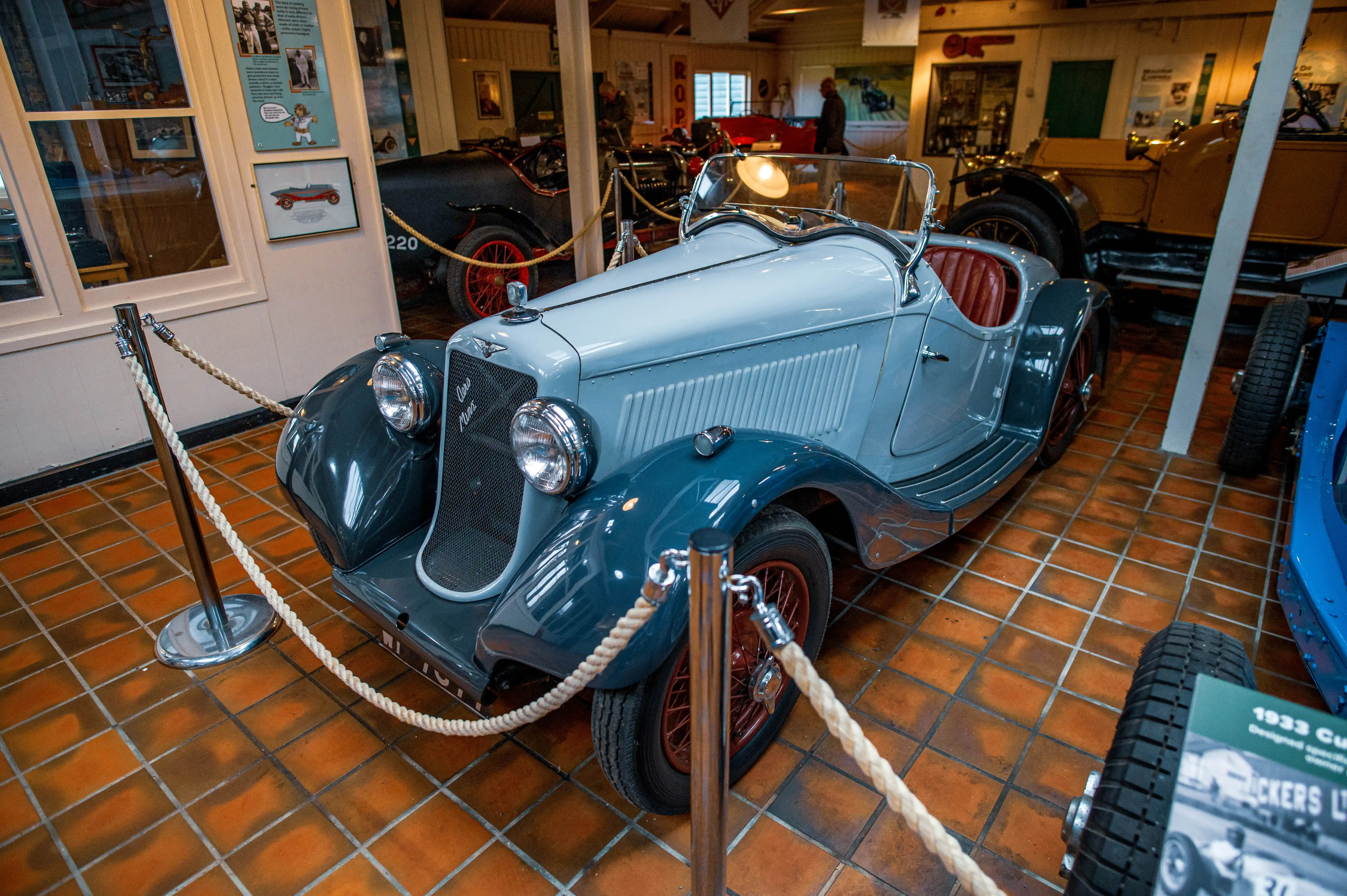 Interior view of a car museum with several vintage automobiles displayed, featuring a grey and white two-seater in the foreground, with other cars in blue, yellow, and green behind it, all separated by rope barriers.