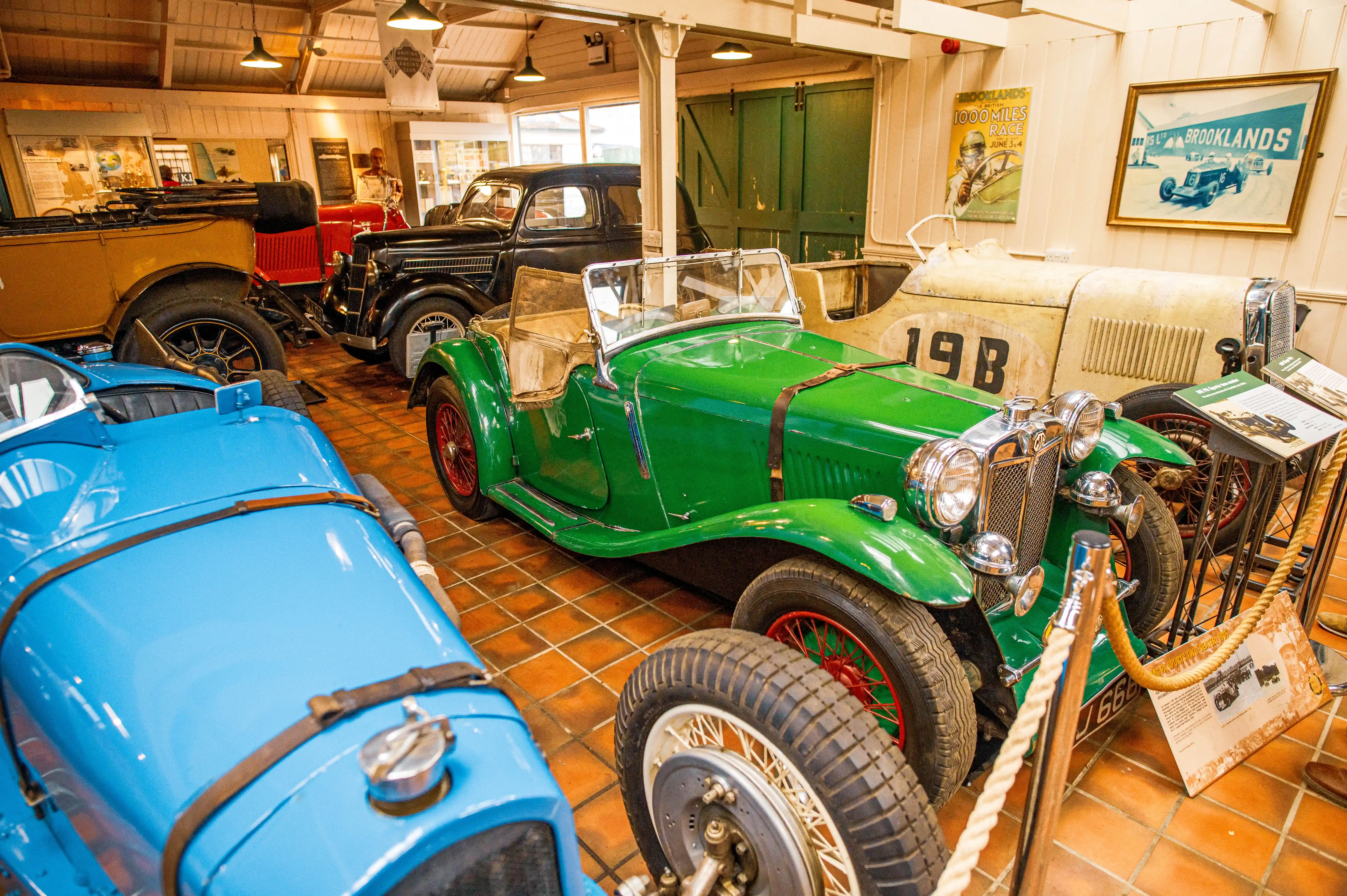 A bright blue 1933 MG K3 racing car with spoked wheels is parked indoors, with a red vintage car partially visible to its left.