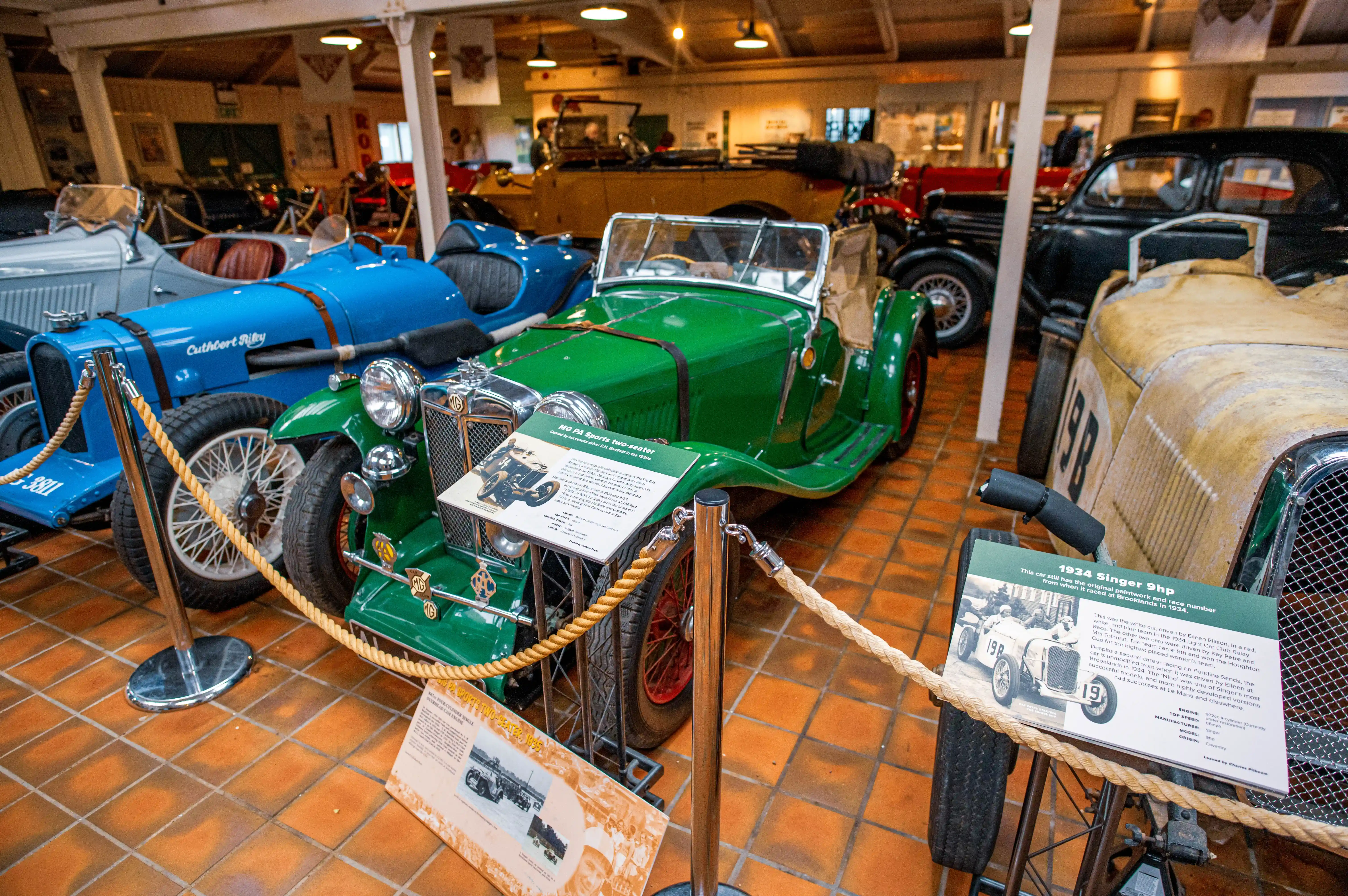 A museum display of vintage cars, featuring a green MG sports car in the foreground with a descriptive plaque, a blue Cuthbert Riley racing car to its left, and other classic vehicles in the background.