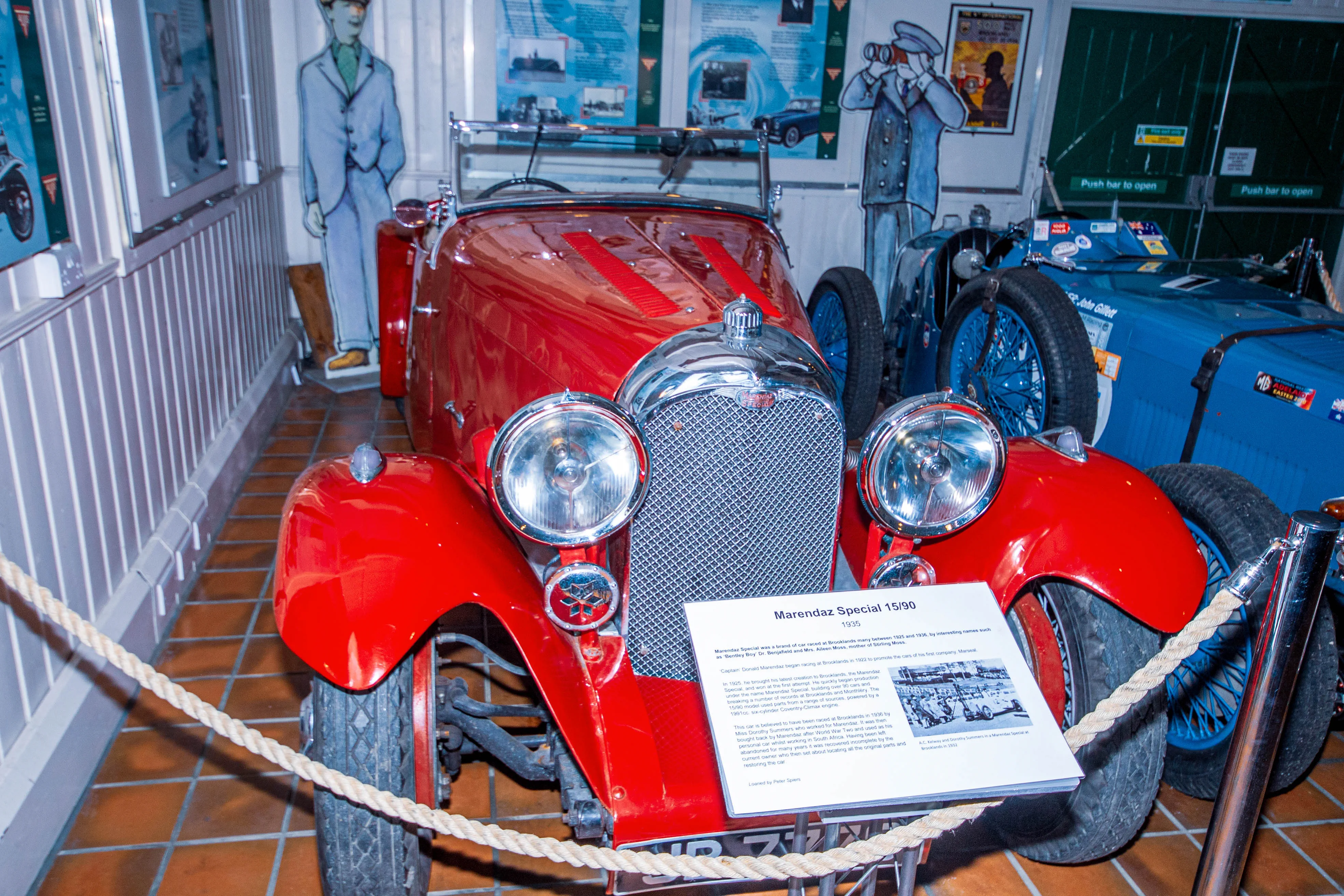 A close-up of a vintage red Marendaz Special 15/90 car from 1935, with its distinctive grille and headlights. A descriptive plaque is placed in front of it. Another blue vintage car is visible to the right.