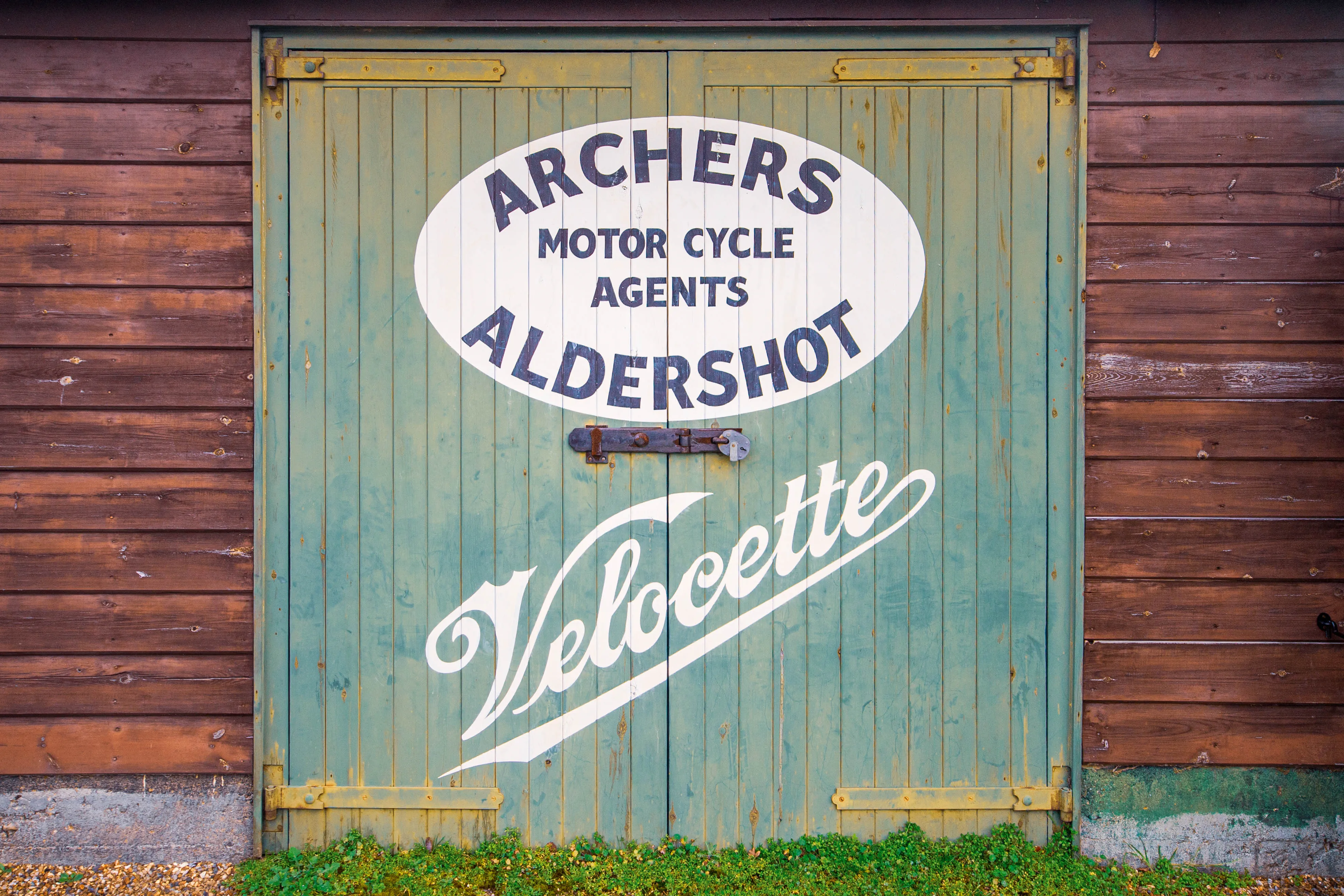 Old green double doors with a painted sign for Archers Motor Cycle Agents in Aldershot and a Velocette logo.