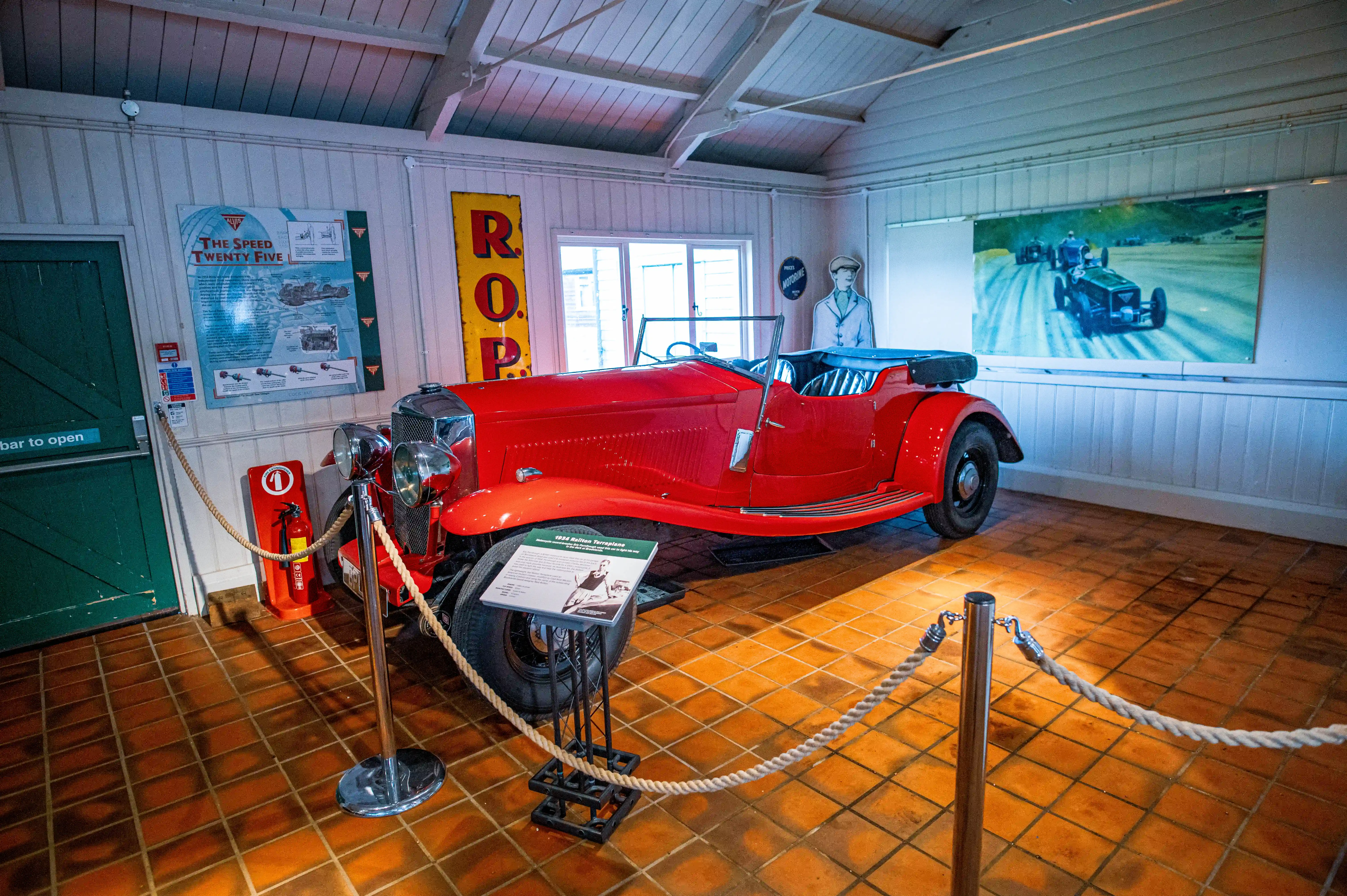 A bright red vintage convertible sports car is displayed indoors on a tiled floor. Walls are decorated with posters and a painting of old race cars. Rope barriers cordon off the exhibit.