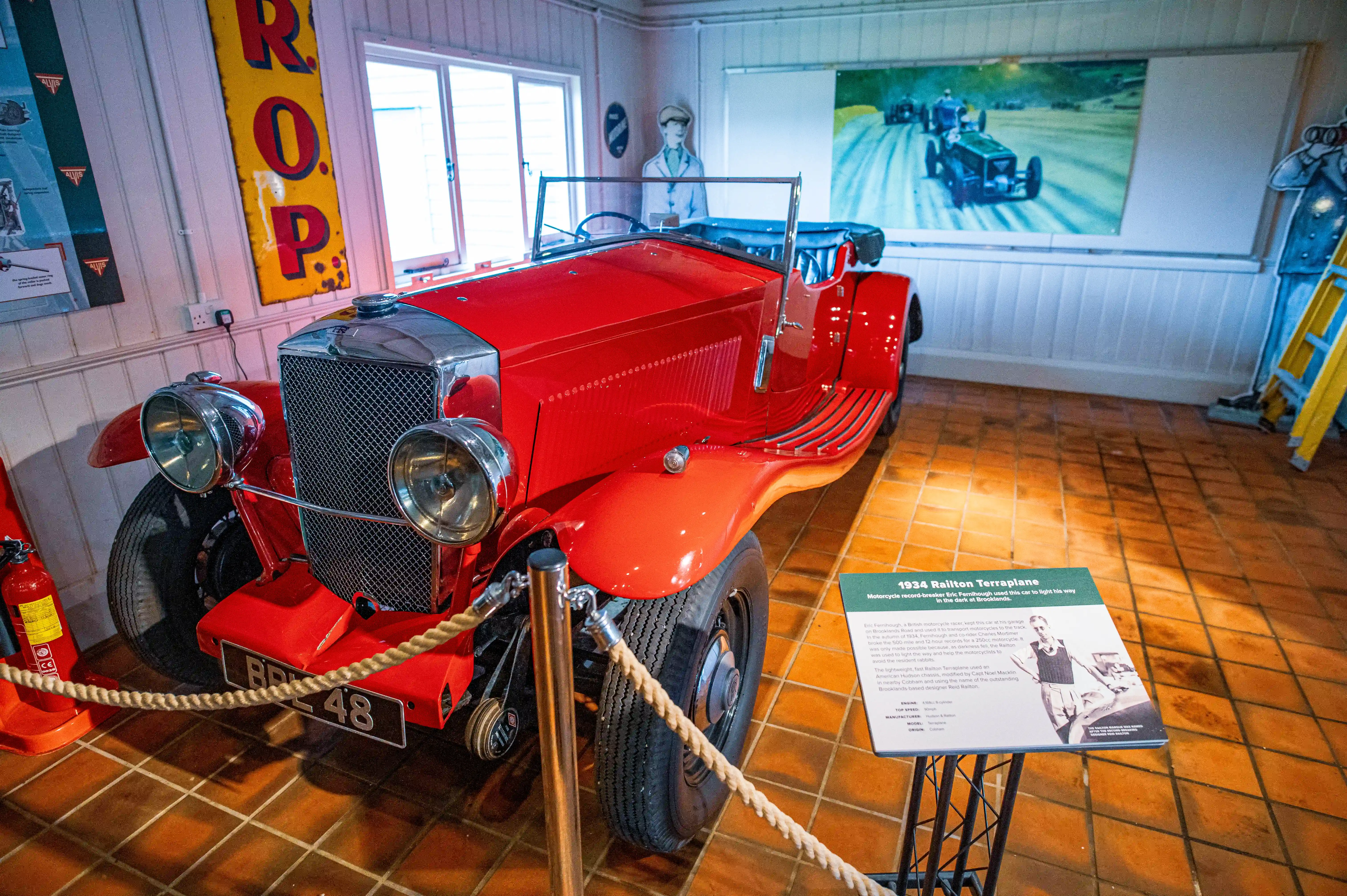A bright red vintage convertible sports car is displayed indoors on a tiled floor. Walls are decorated with posters and a painting of old race cars. Rope barriers cordon off the exhibit.