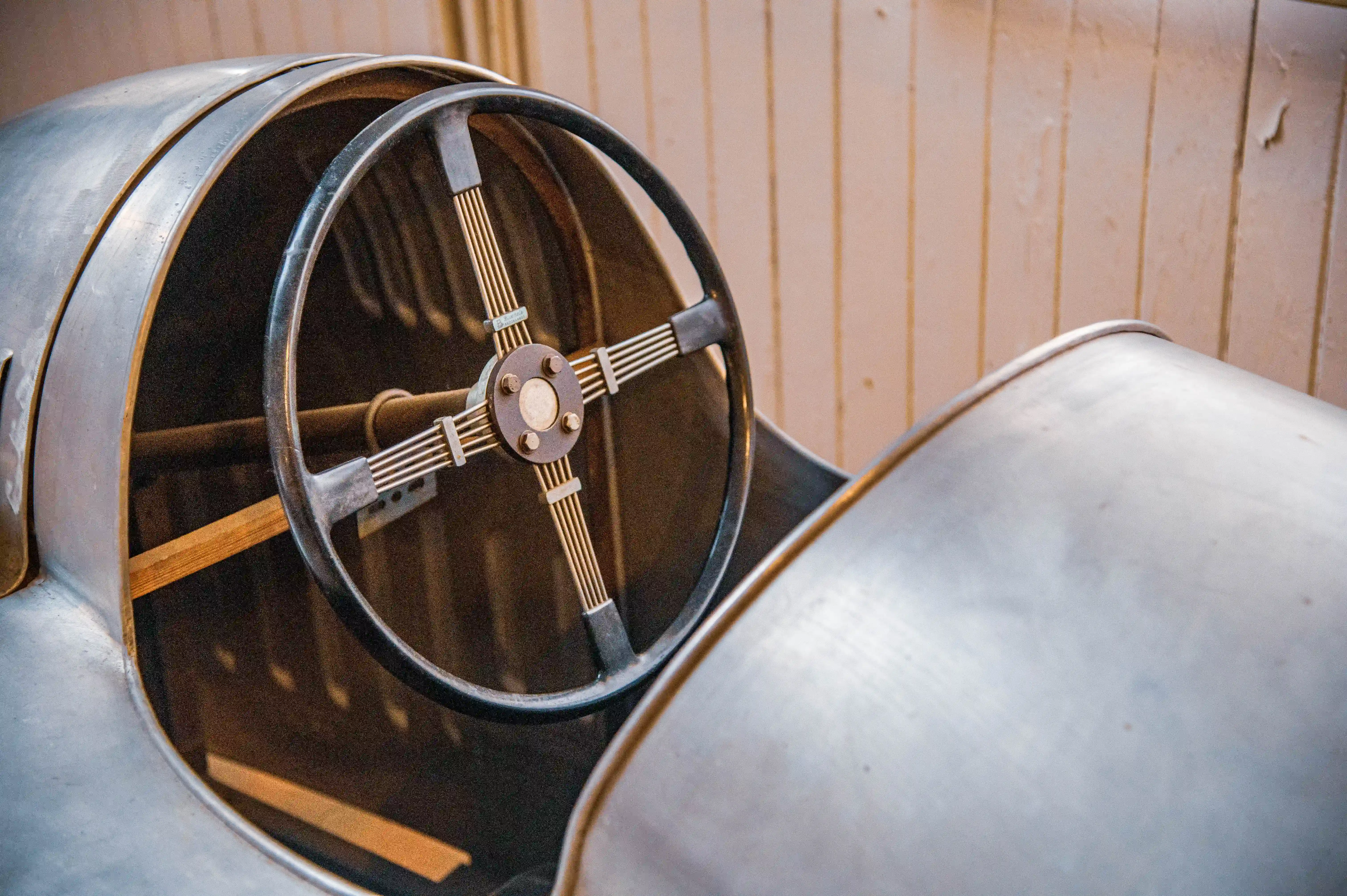 Close-up of a vintage car's steering wheel, showing the dark rim, metallic spokes, and central hub. The polished metal body of the car is visible to the left and right, with a wooden dashboard element behind the wheel. The background consists of vertical wooden panelling.