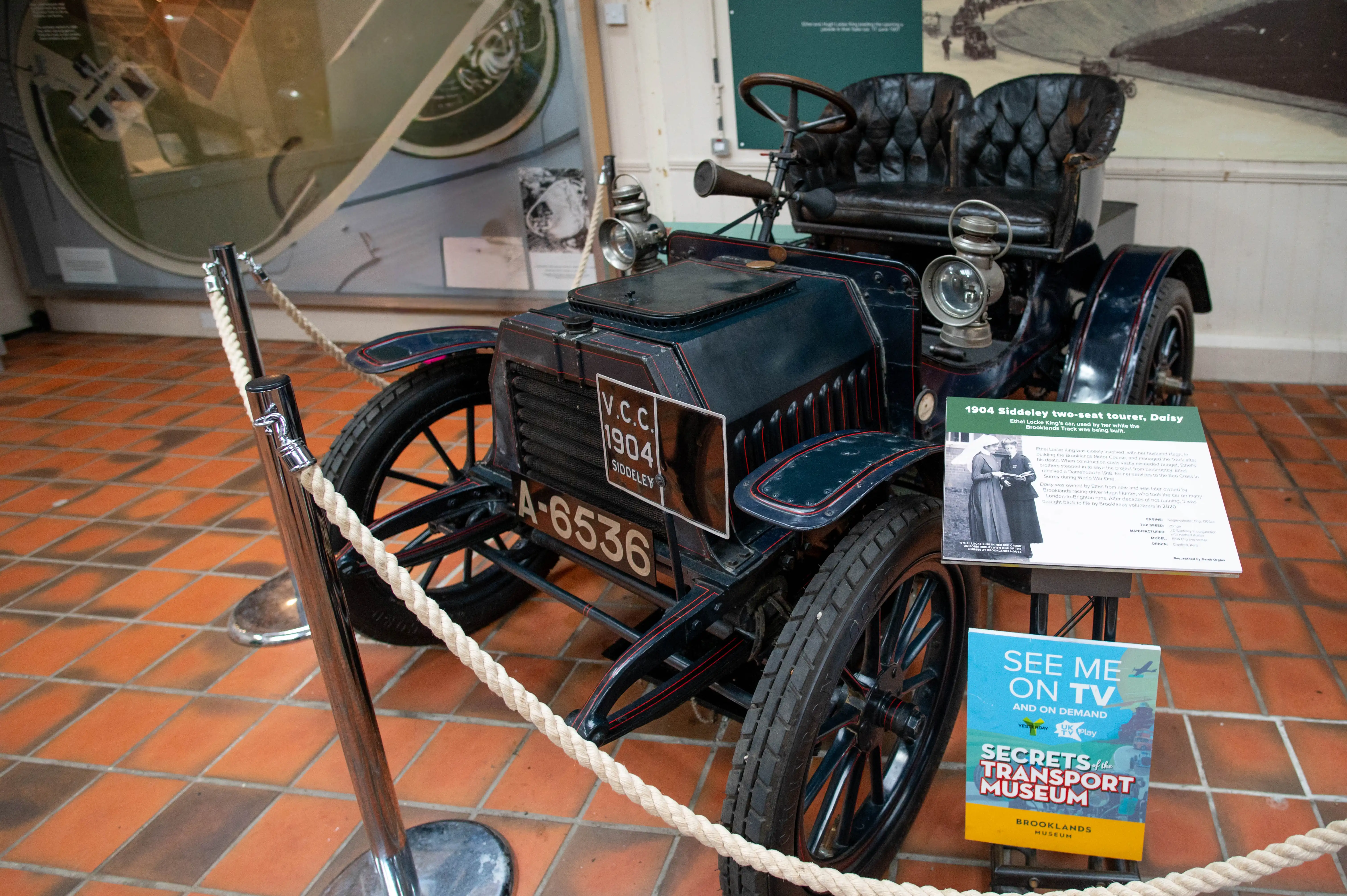 A dark blue 1904 Siddeley two-seat tourer with large spoked wheels and leather seats is on display in a museum. A rope barrier is in front of the car. A sign next to the car provides information about its history and an advertisement for Secrets of the Transport Museum.