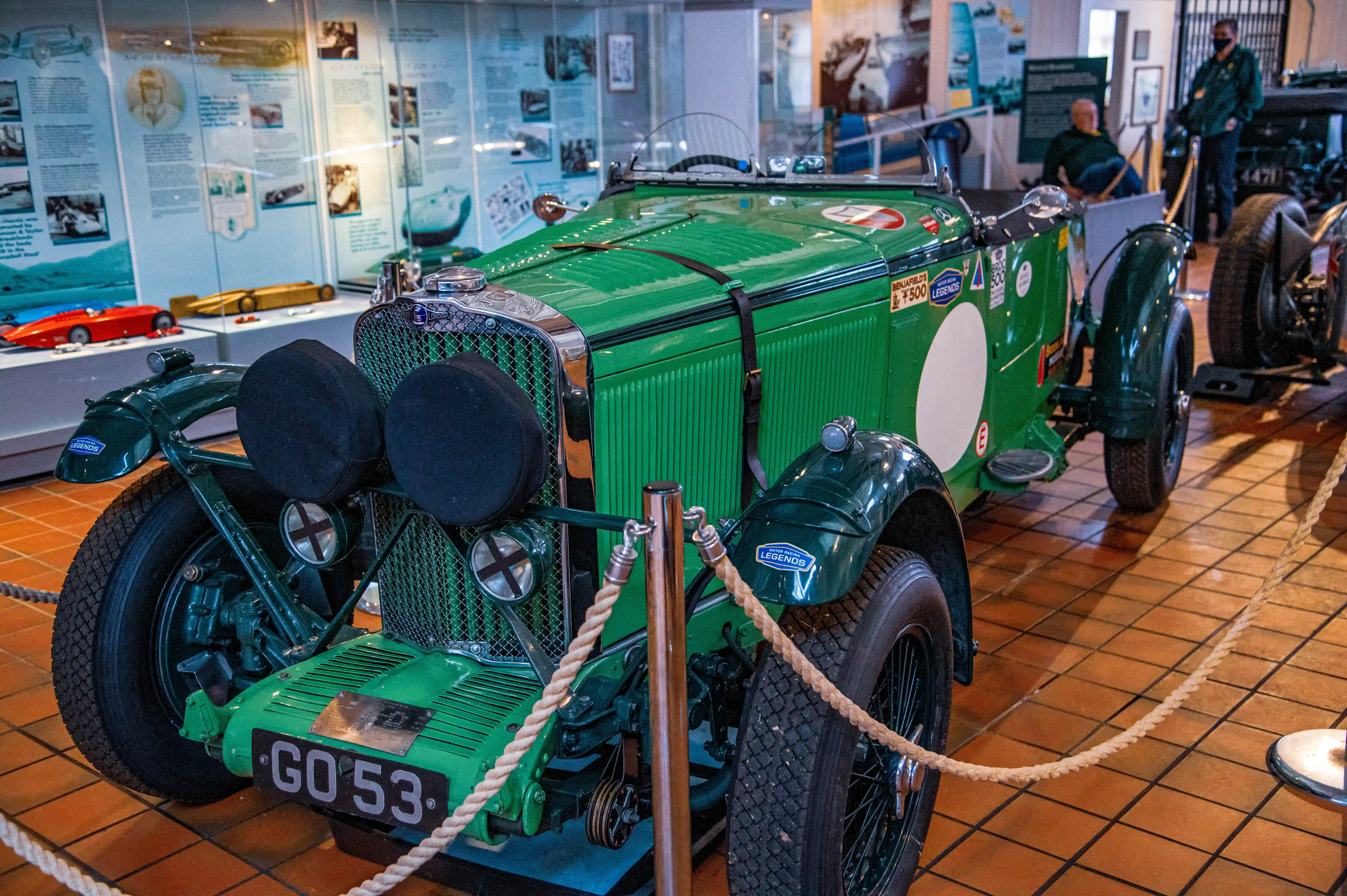 A green, open-top vintage racing car with the registration plate 'GO 53' is parked indoors. The car has large, dark circular headlight covers and is displayed behind a rope barrier. In the background, museum exhibits with historical images and other vehicles are visible.