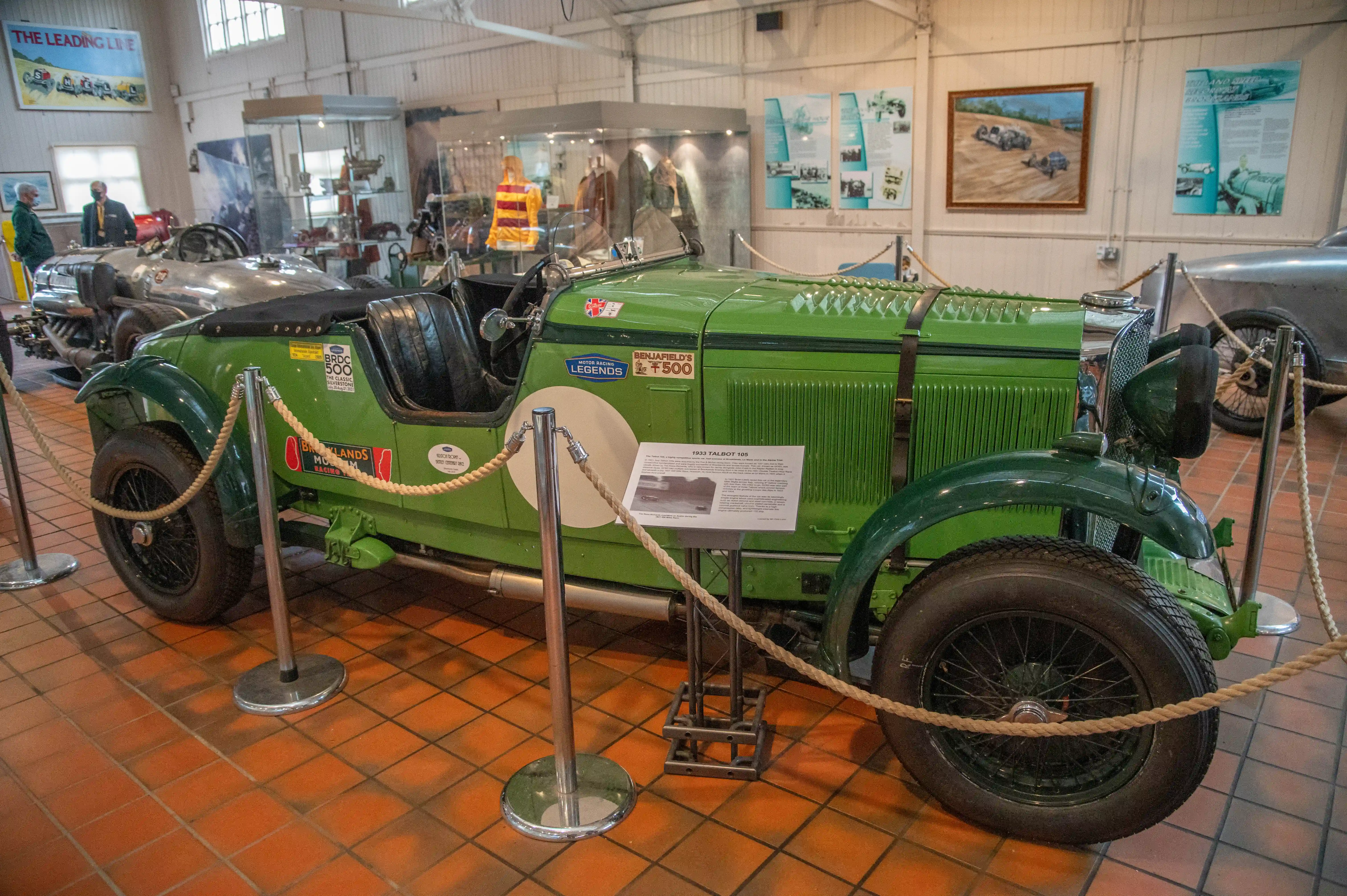 A vintage green Talbot 105 sports car with spoked wheels and a black soft-top is displayed in a museum. Information plaques and ropes are present around the car. In the background, other vehicles, posters, and display cases with racing memorabilia are visible.