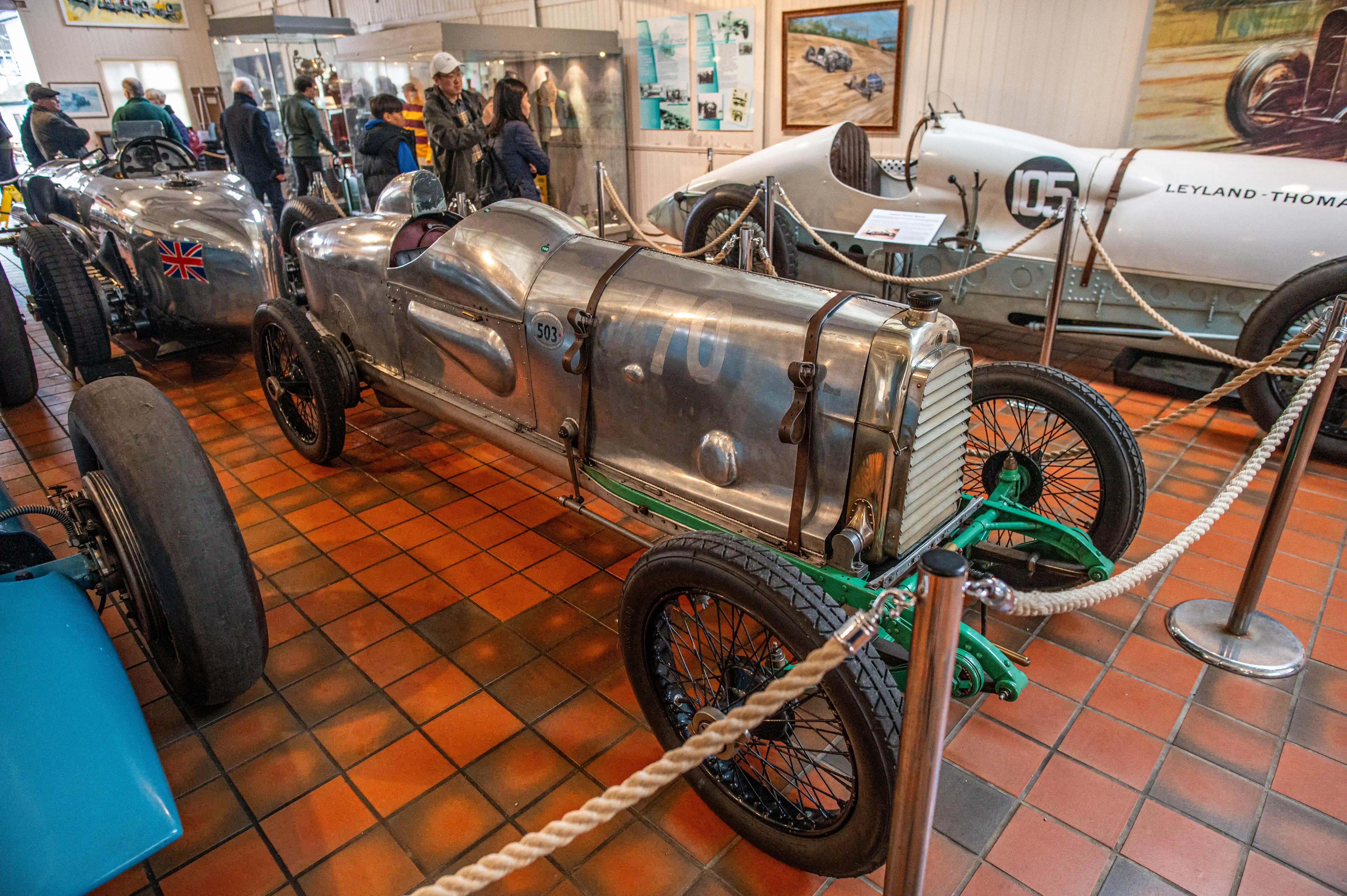 A vintage silver racing car with the number 270 on its body, displayed in a museum with ropes in the foreground and other cars in the background.
