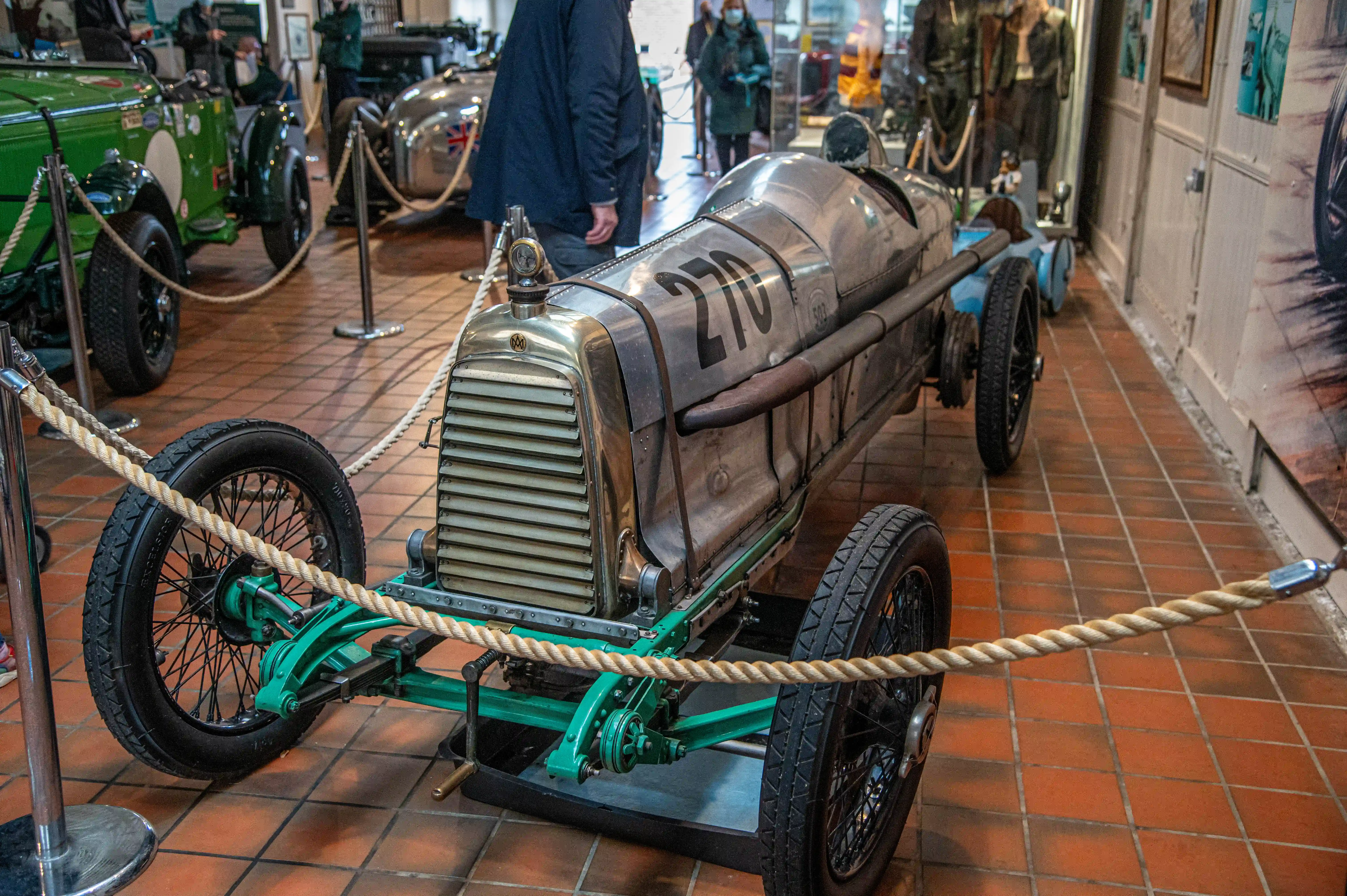 Two vintage racing cars, one silver with the number 70 and leather straps, and another silver car with a Union Jack flag, are parked indoors on a tiled floor. A white racing car with the number 105 and the text 