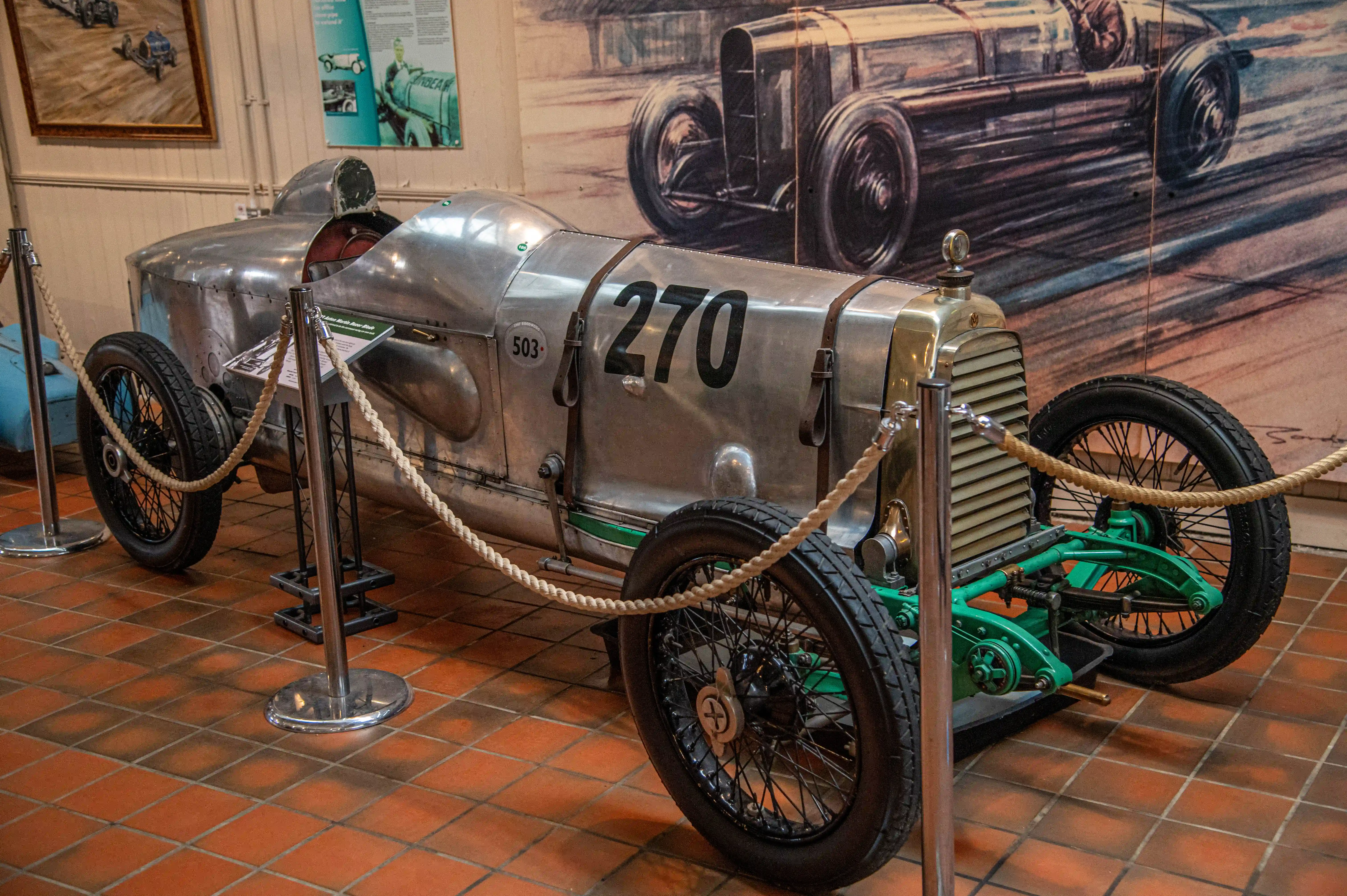 Two vintage racing cars, one silver with the number 70 and leather straps, and another silver car with a Union Jack flag, are parked indoors on a tiled floor. A white racing car with the number 105 and the text 