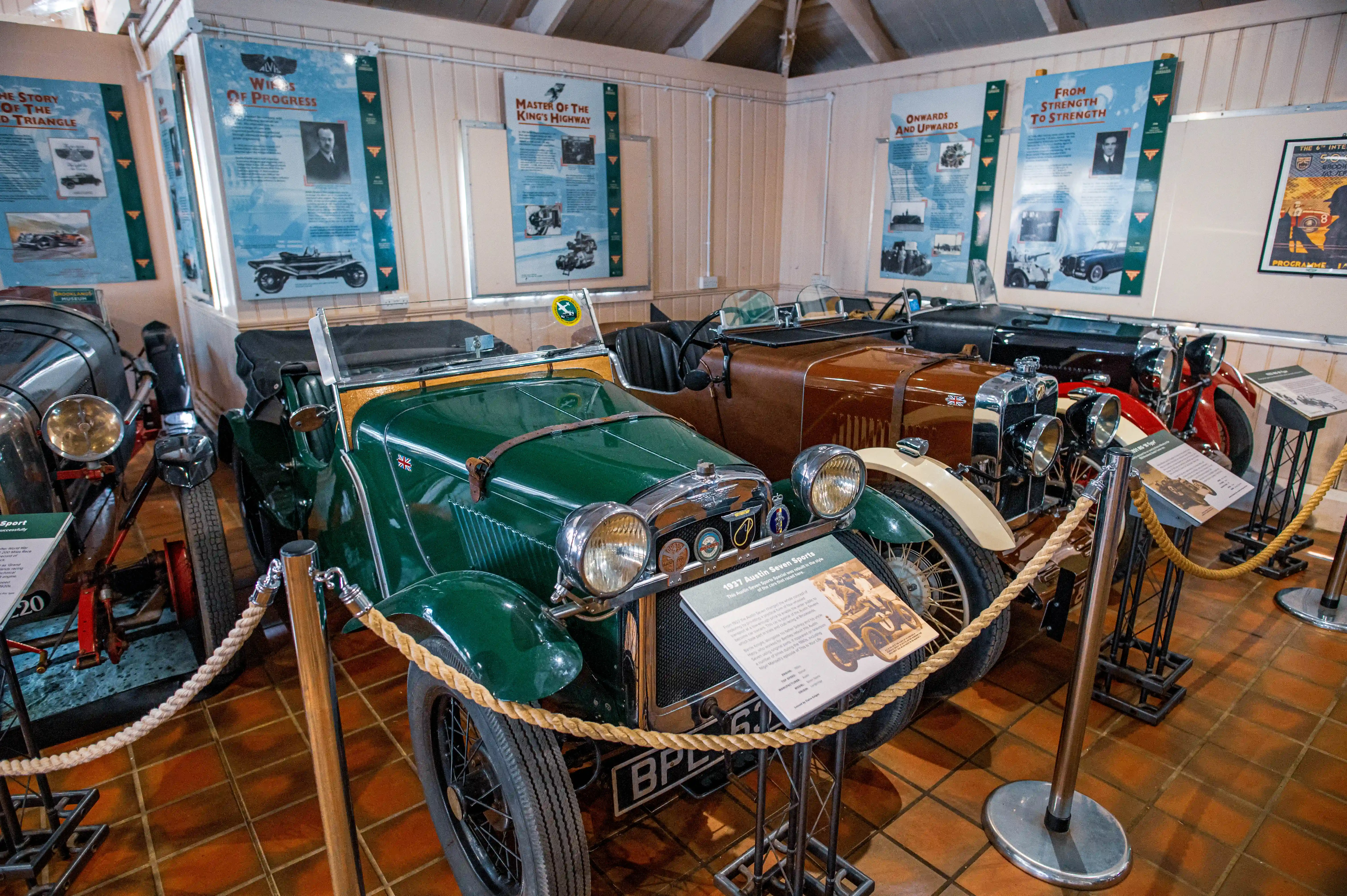 A 1937 green Austin Seven Sports car is parked in a museum display, with other vintage cars behind it. Informational posters are mounted on the wall.
