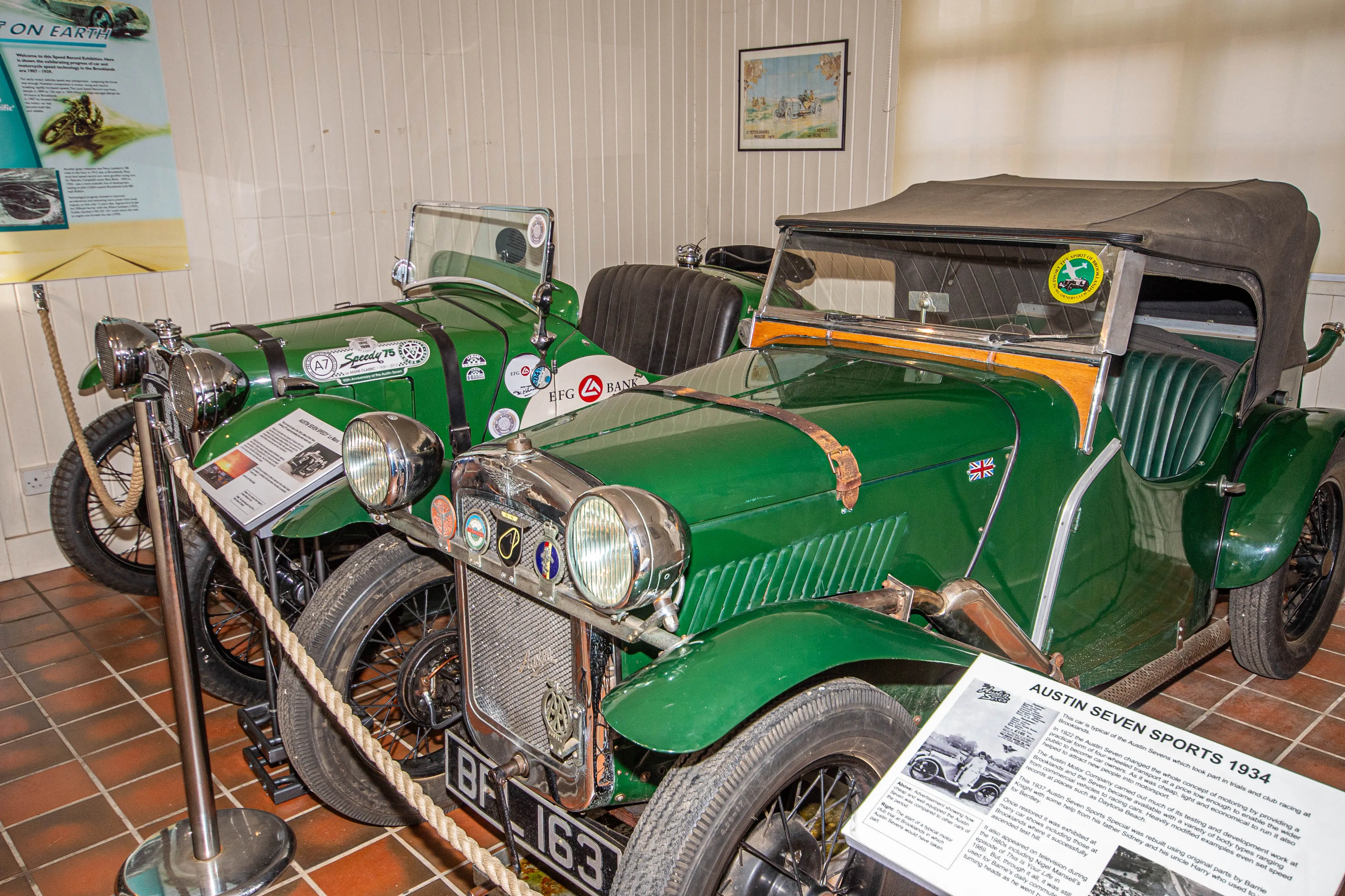 A close-up view of two vintage green Austin Seven Sports cars, one with licence plate 'BF 163', on display in a museum. Informative plaques are placed in front of the cars, and stanchions with ropes separate them from visitors.