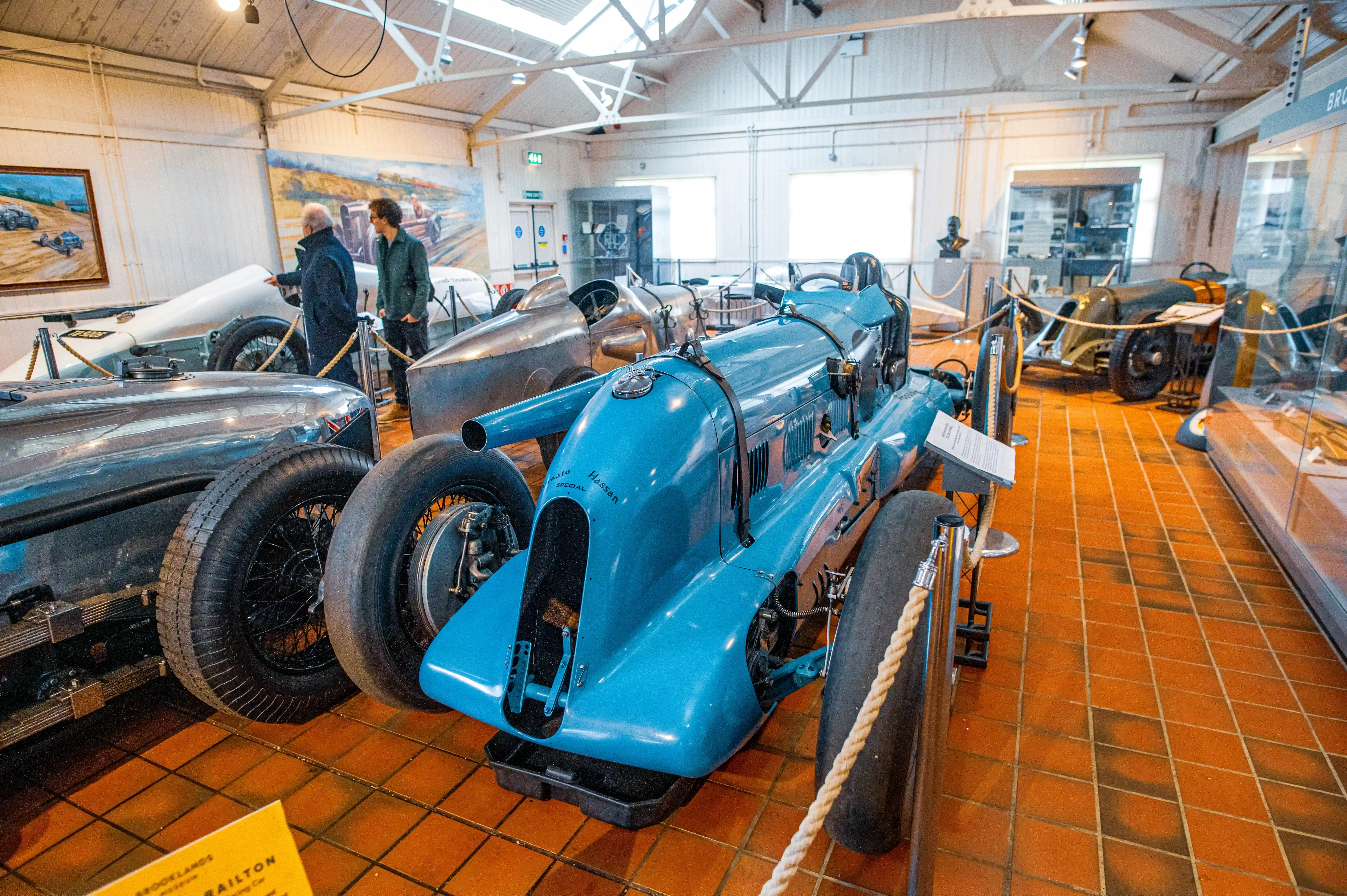 A museum interior displaying vintage racing cars. A bright blue car with Mato Special Hassan written on it is in the foreground, with two people looking at other cars in the background.