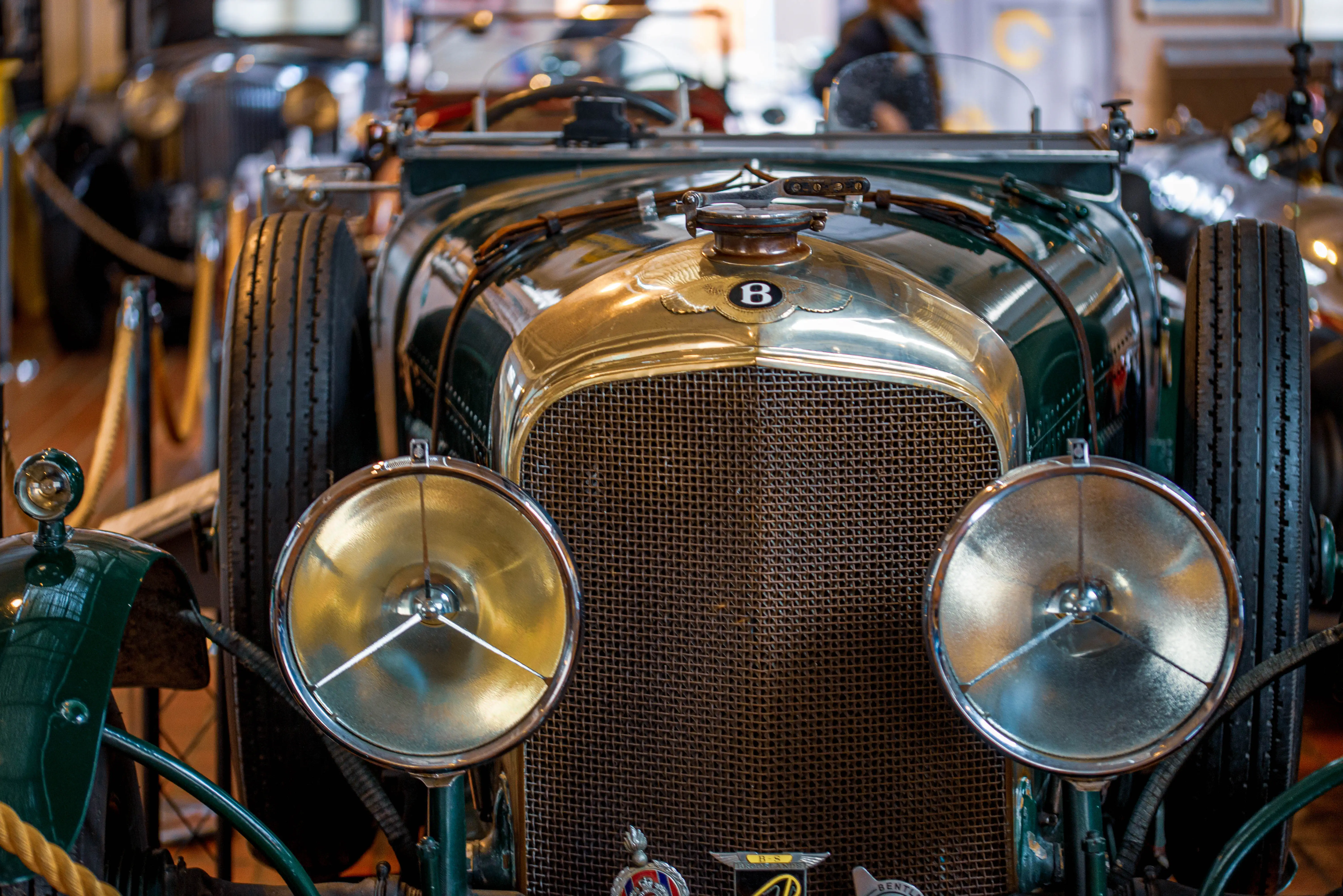 Close-up frontal view of a classic dark green Bentley car, focusing on its brass grille, circular chrome headlights, and the distinctive Bentley 'B' badge on the bonnet.