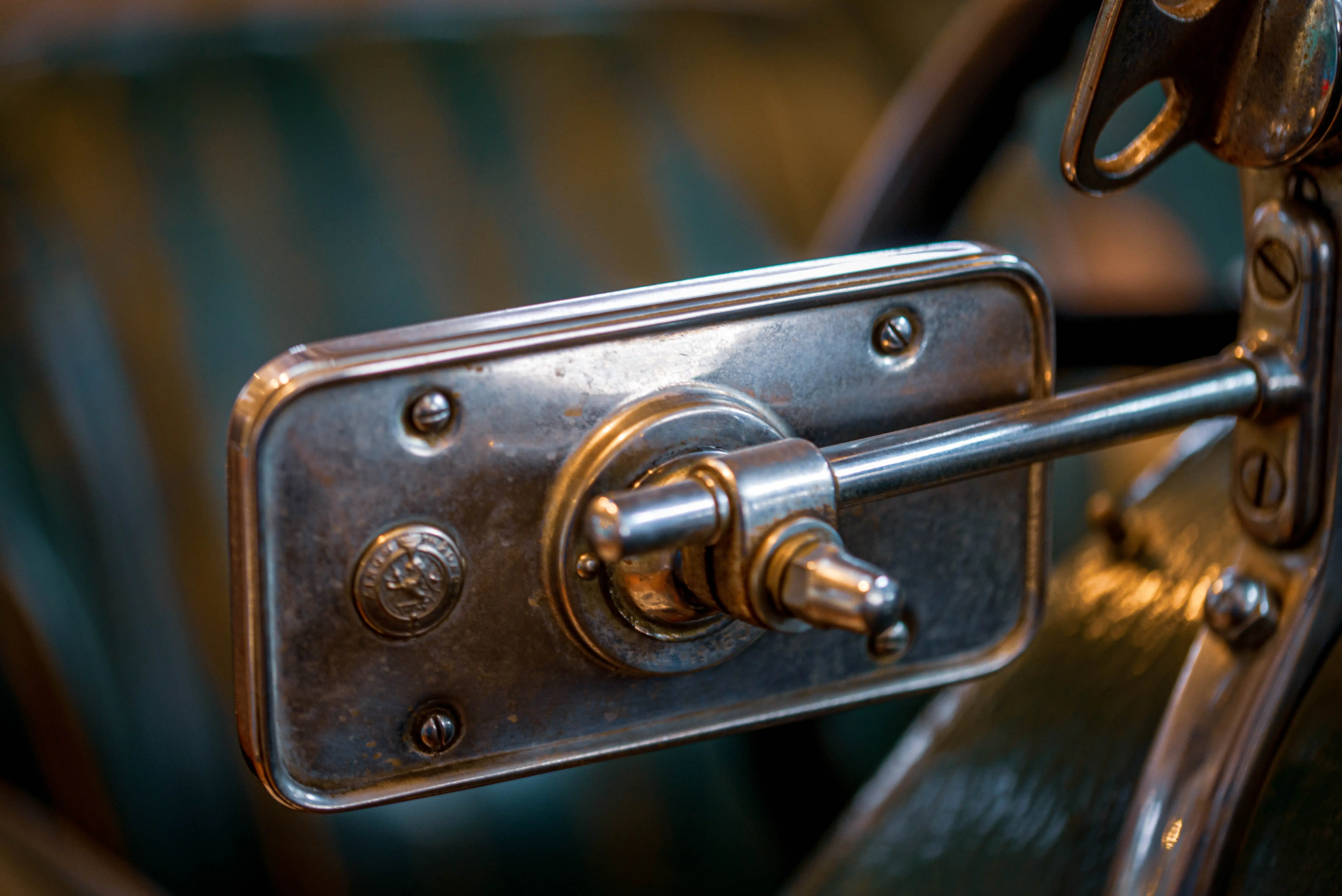 Close-up of a vintage car's wing mirror adjustment lever and housing, showing detailed metalwork and screws.