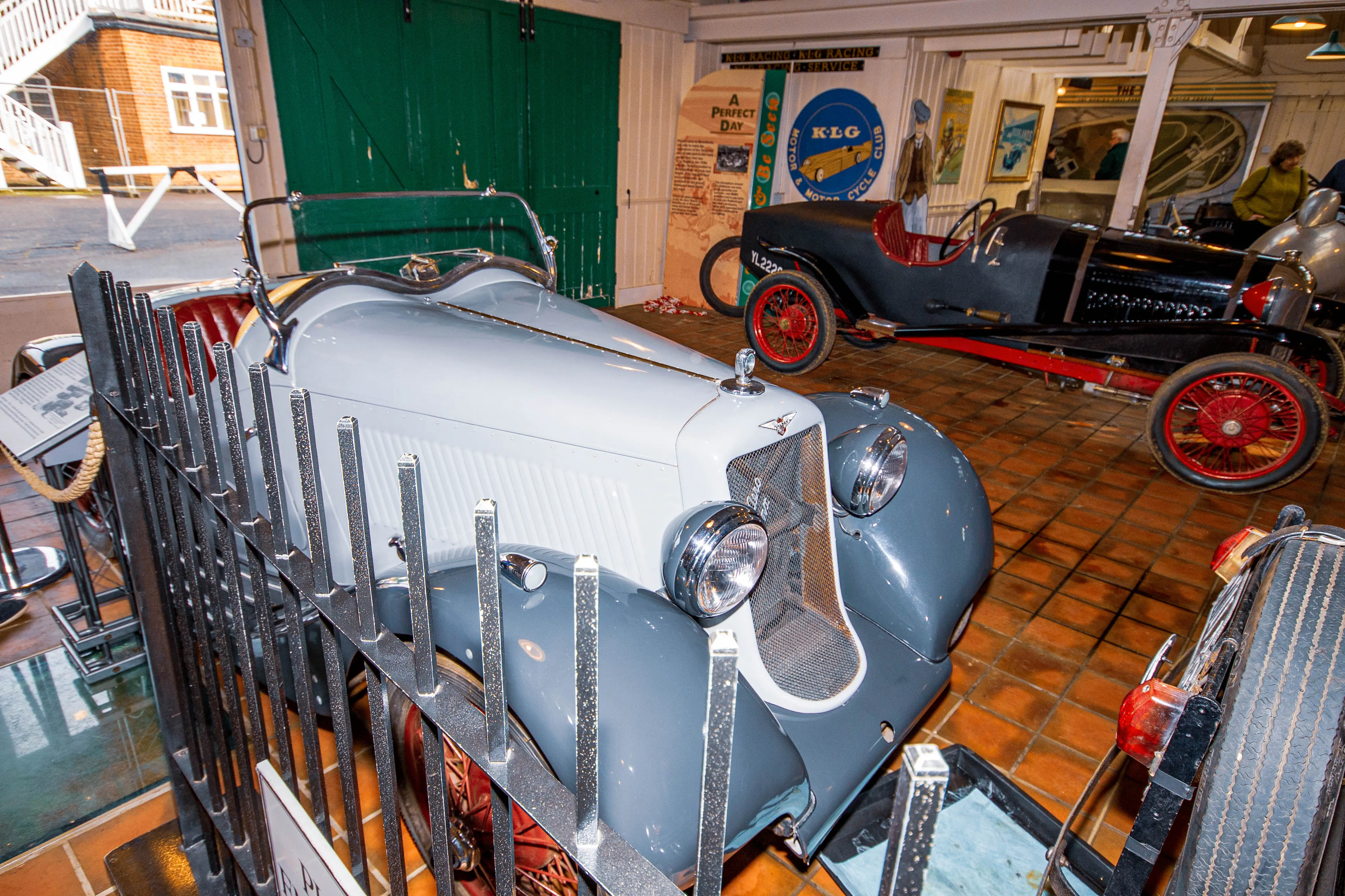 Close-up of the front of a vintage light grey convertible car, showing its chrome grille, headlights, and red interior, with a metal fence in the foreground and another vintage car in the background.